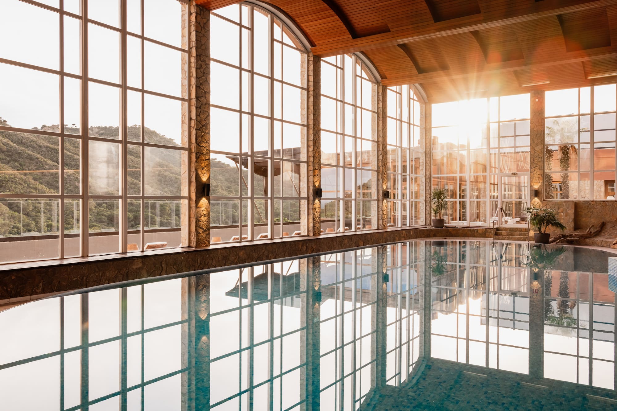 Sunlit indoor pool with panoramic mountain views and arched windows.
