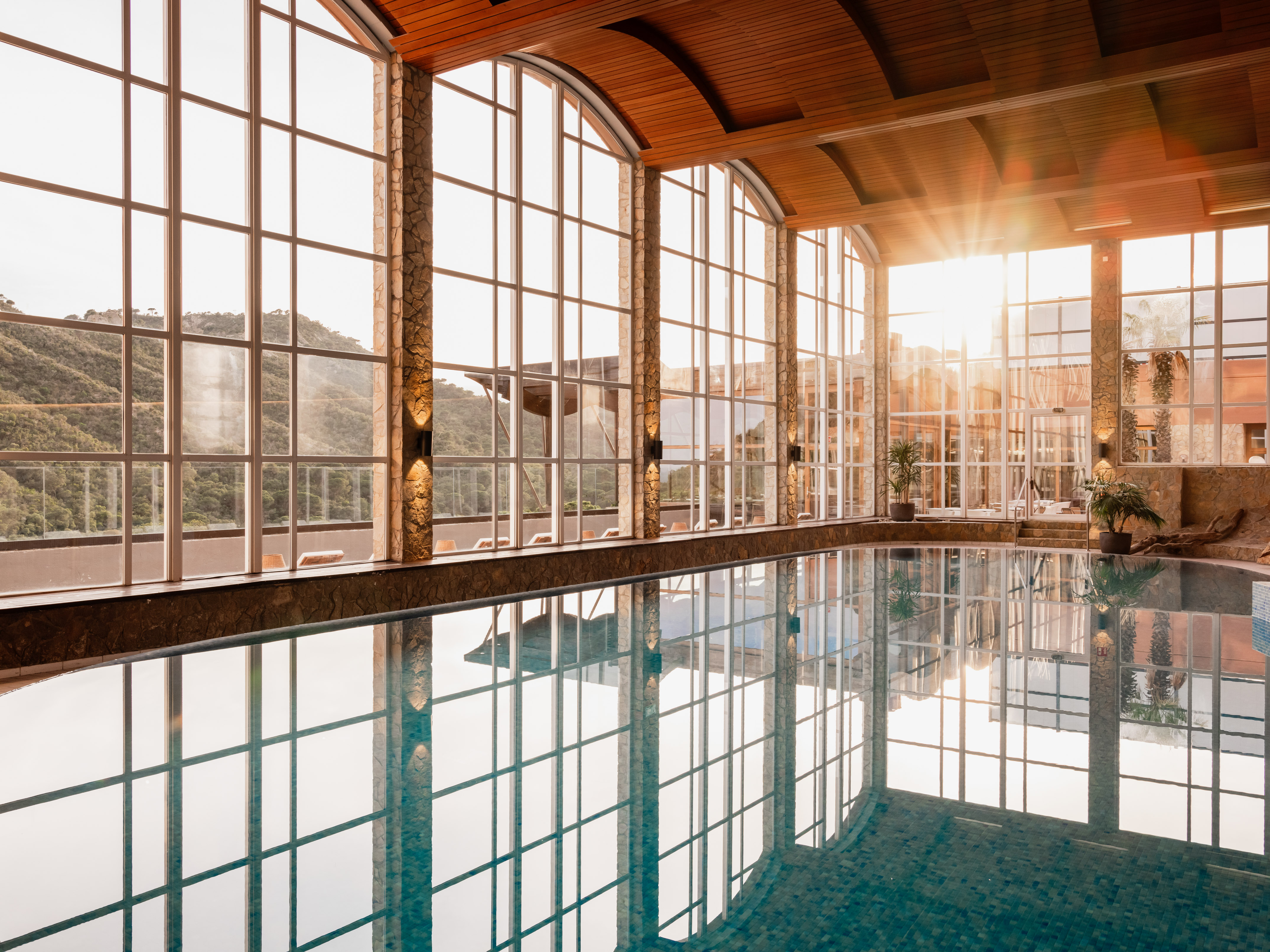 Sunlit indoor pool with panoramic mountain views and arched windows.