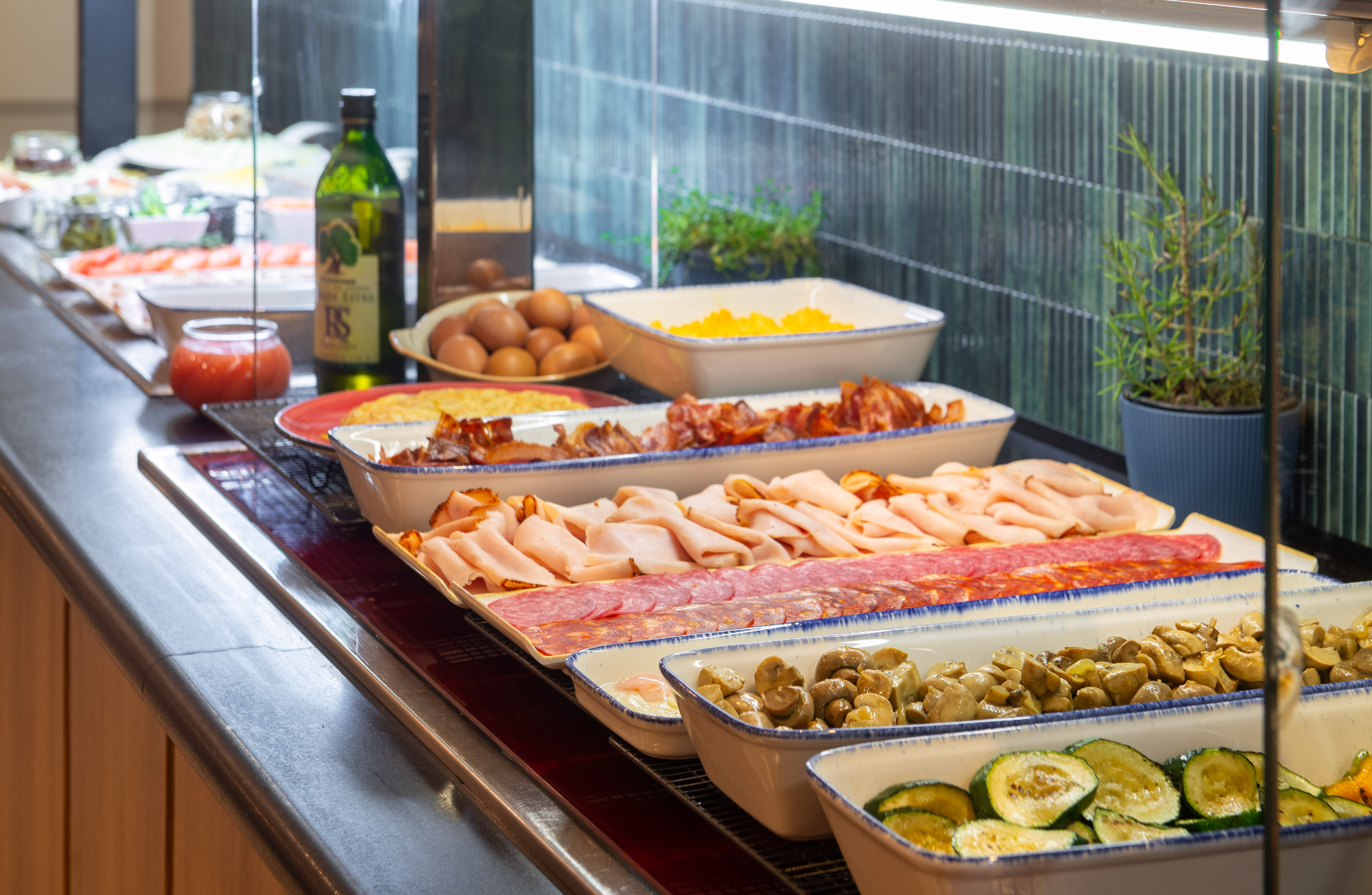 Breakfast buffet with cold cuts, crispy bacon, eggs, grilled zucchini, and mushrooms.