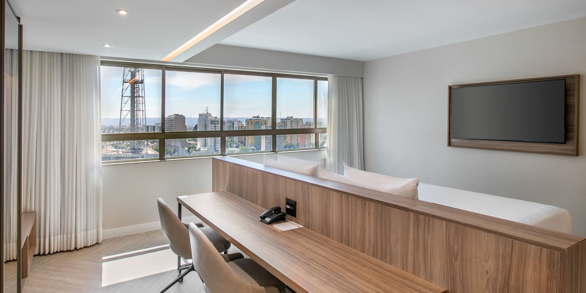 Modern hotel suite with city skyline view, workspace, and flat-screen TV.