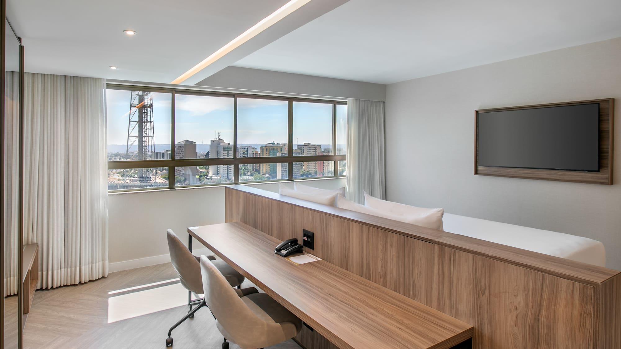 Modern hotel suite with city skyline view, workspace, and flat-screen TV.
