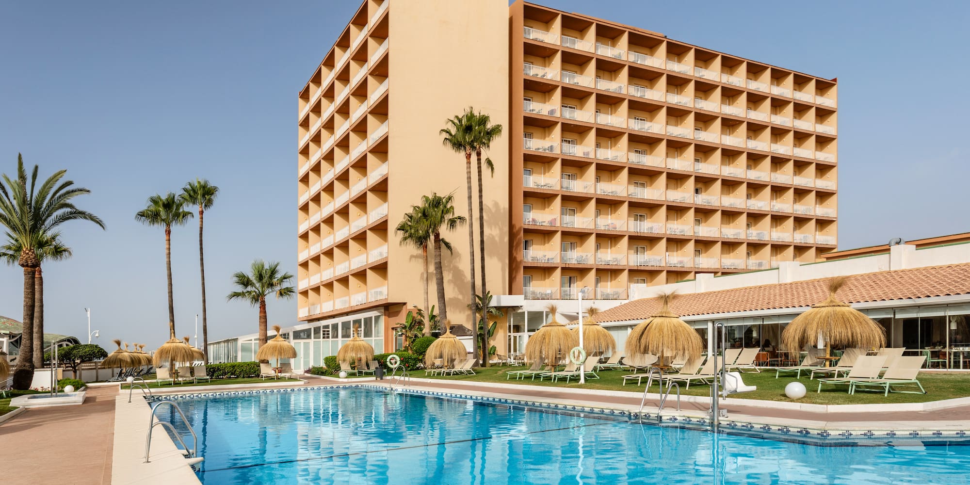 Sunny resort hotel with blue swimming pool, palm trees, and balconies.