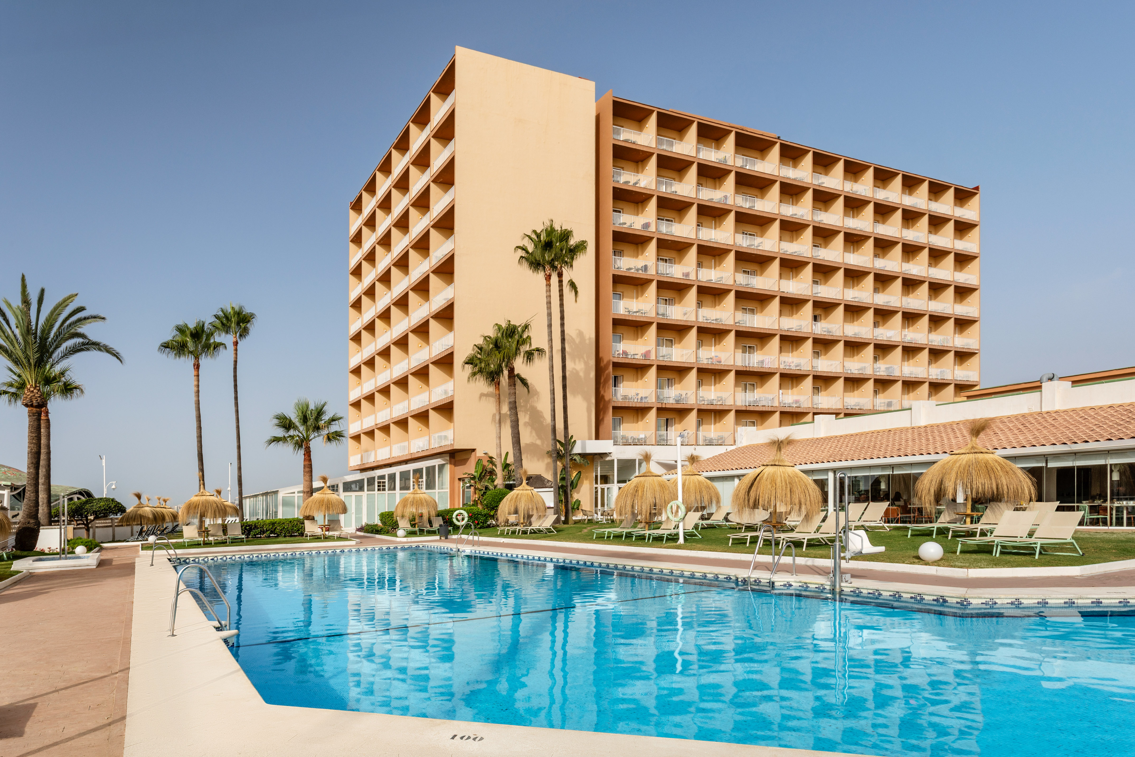 Tan hotel building with balconies, swimming pool, lounge chairs, and palm trees.