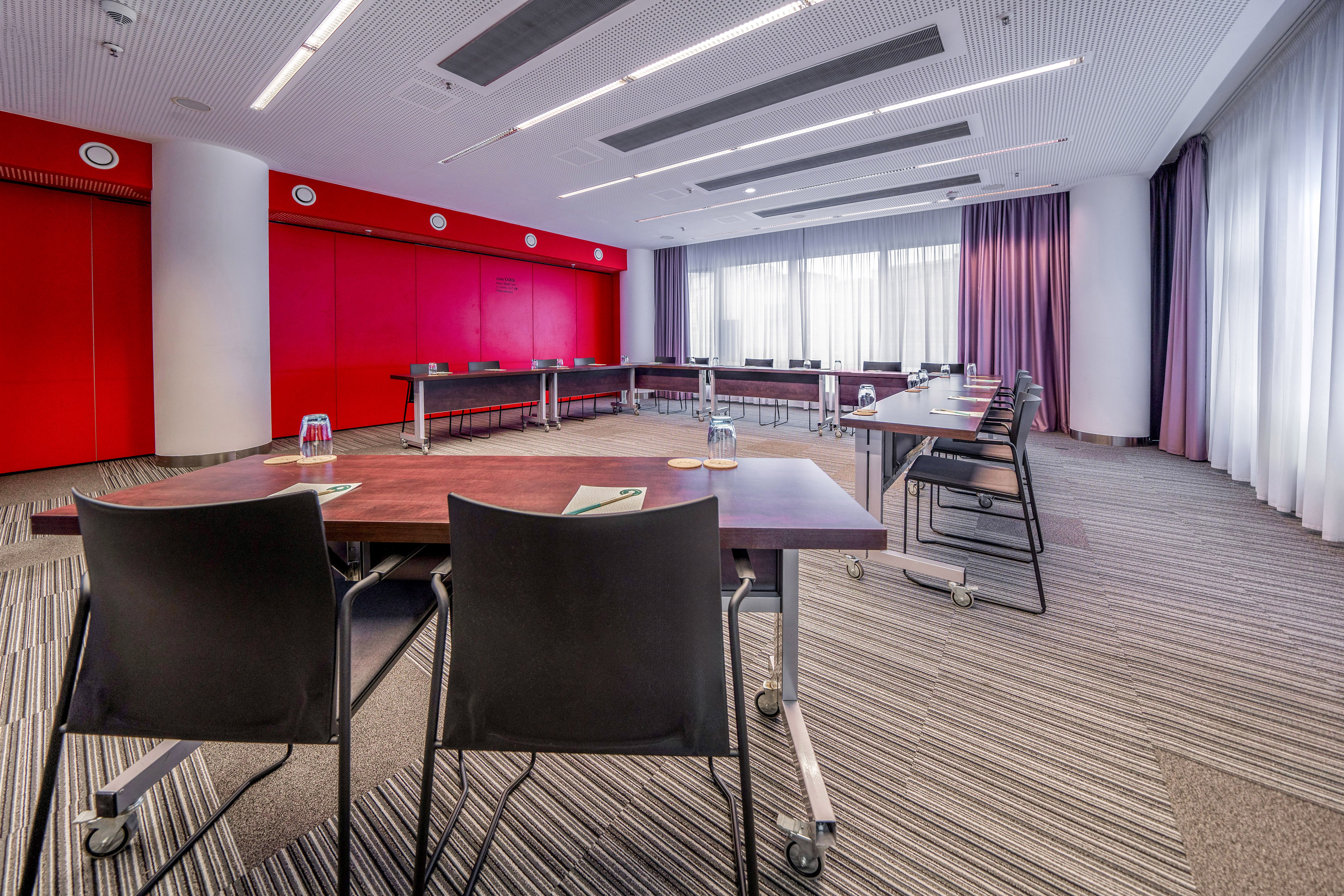 Modern conference room with U-shaped tables, red accent wall, and large windows.