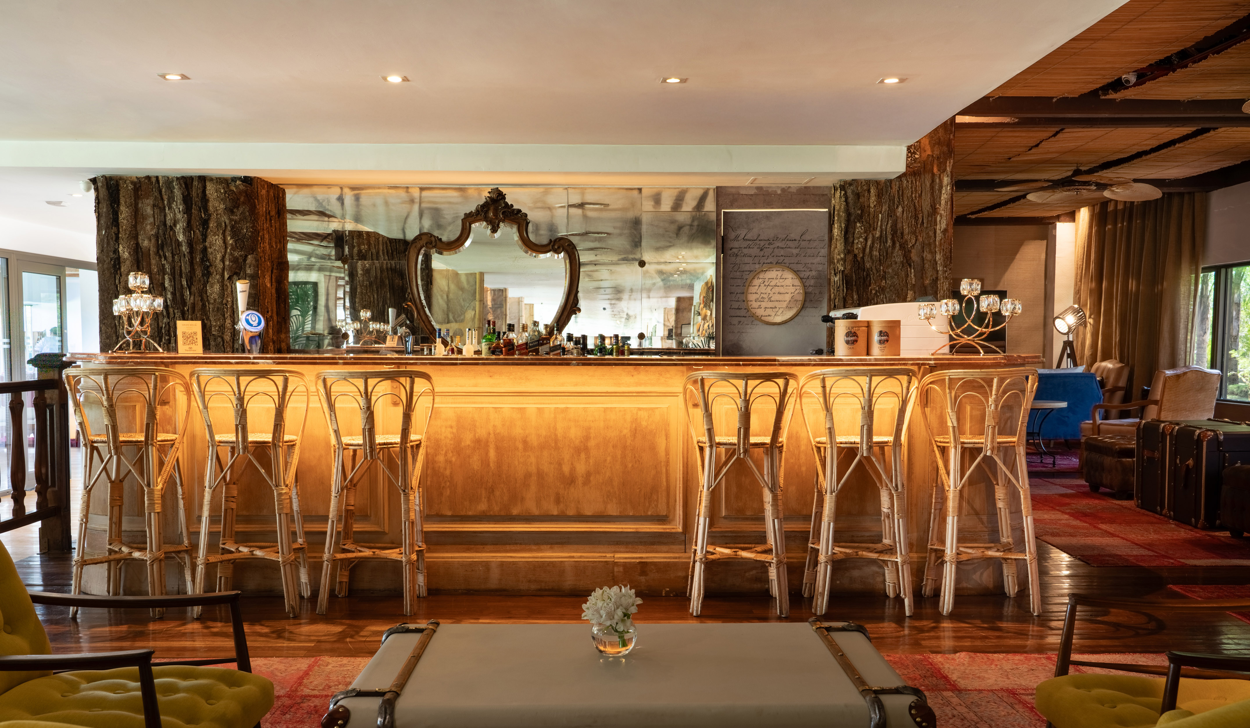 Rustic hotel bar with wooden counter, rattan stools, and mirrored wall.