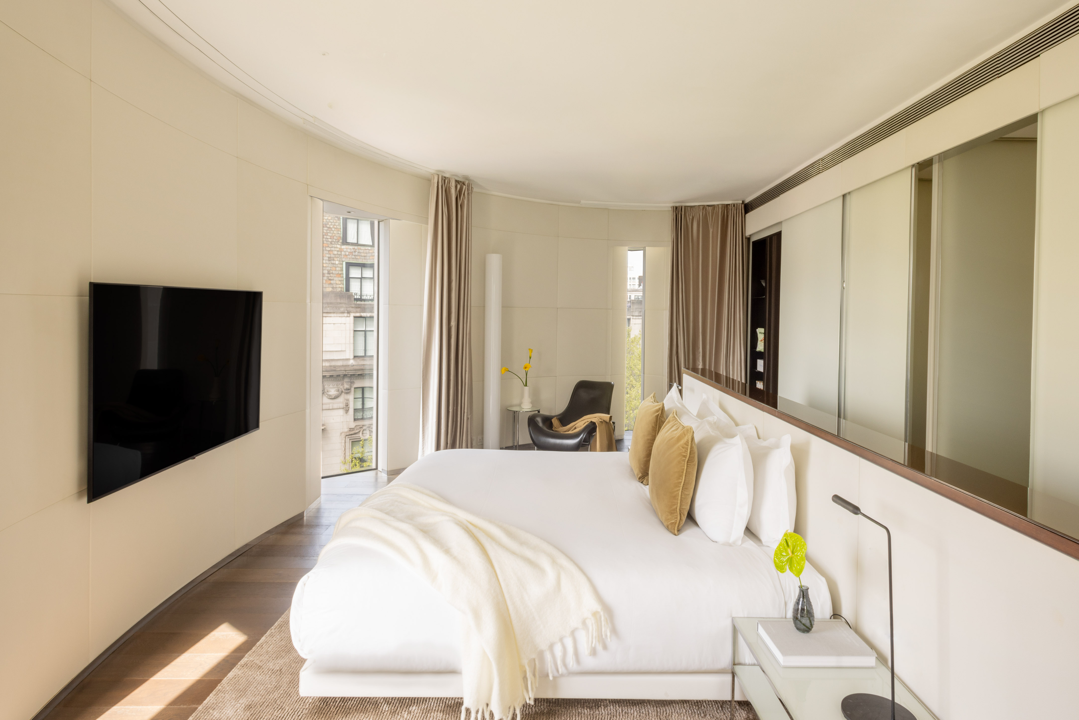 Modern luxury bedroom with curved walls, white bed, large windows, city view.
