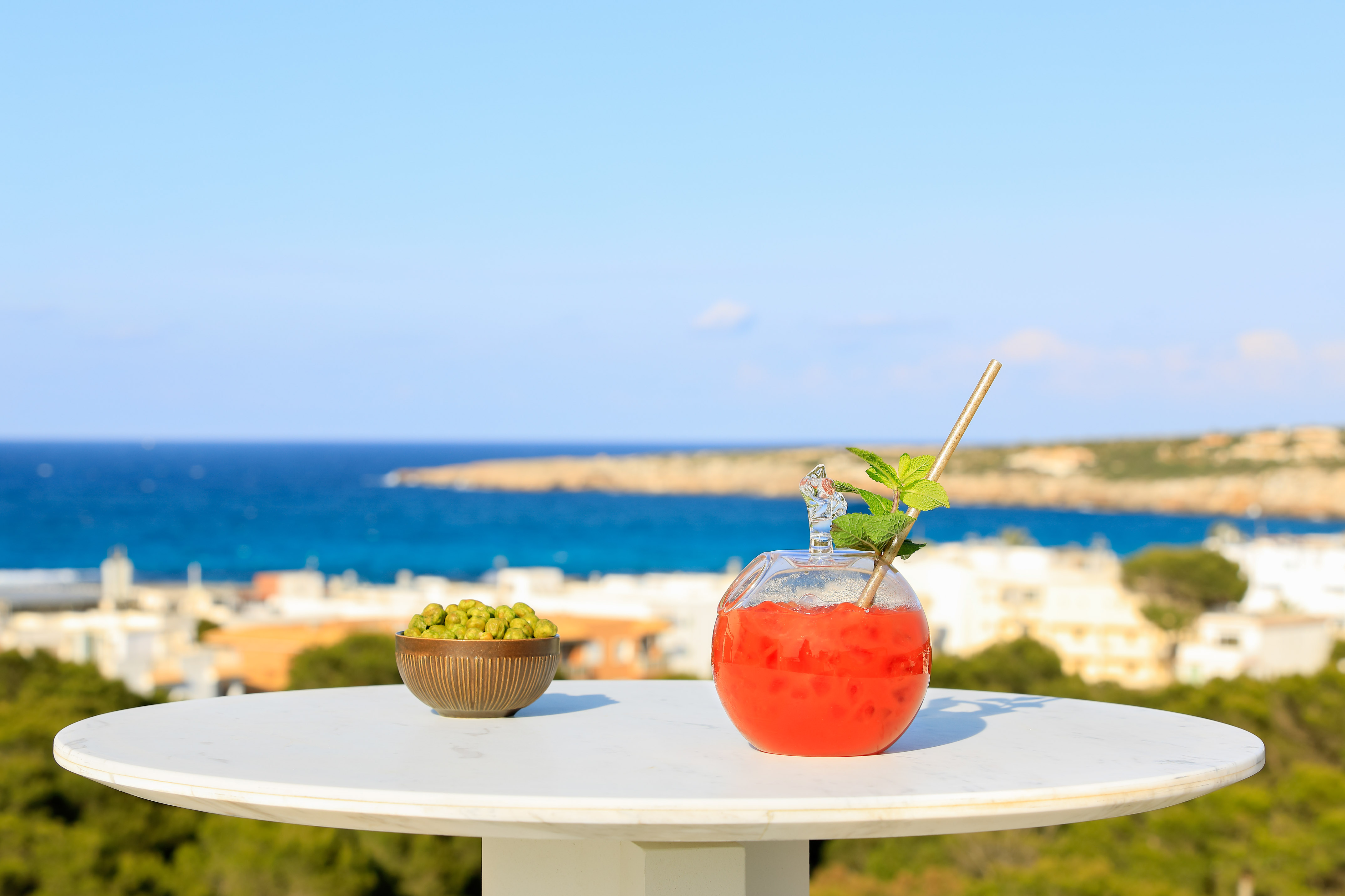 Tropical red cocktail with mint, green snacks on table, ocean view.