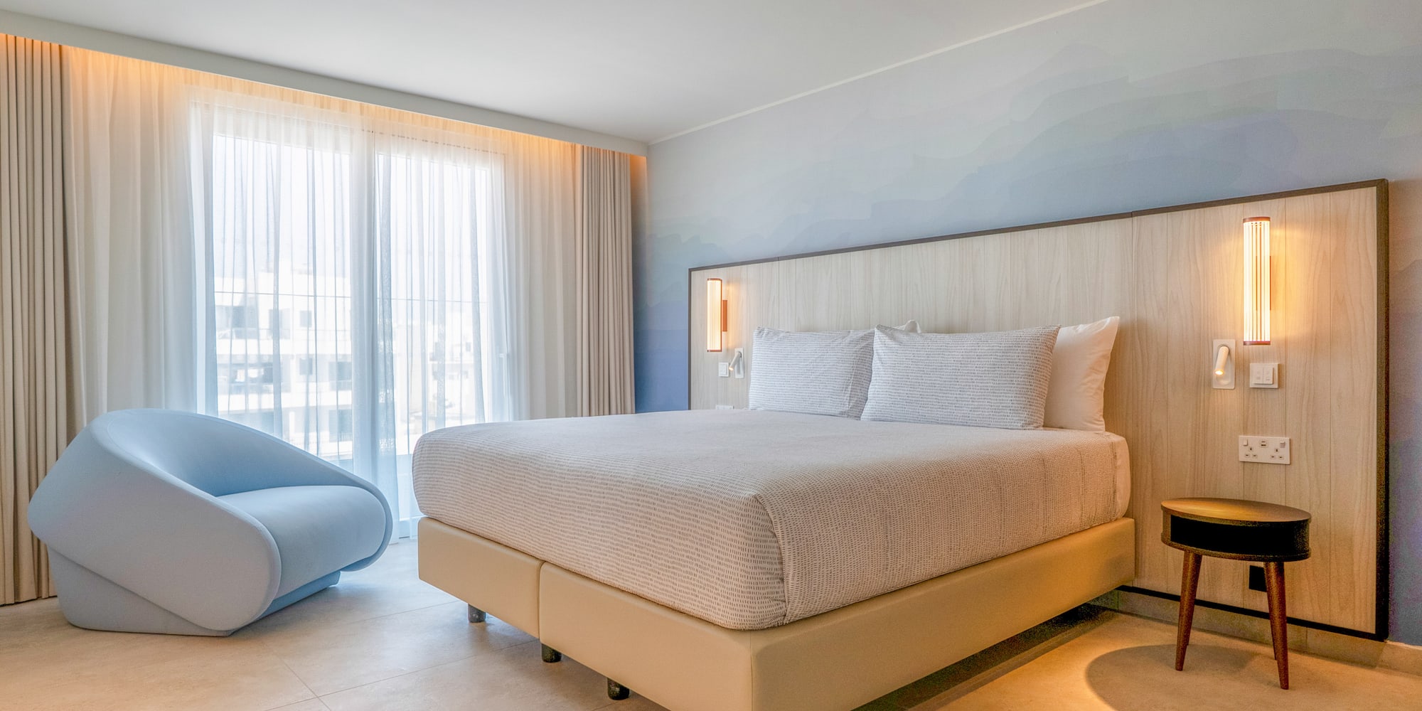 Modern hotel bedroom with king bed, light wood headboard, blue accent chair, large window.