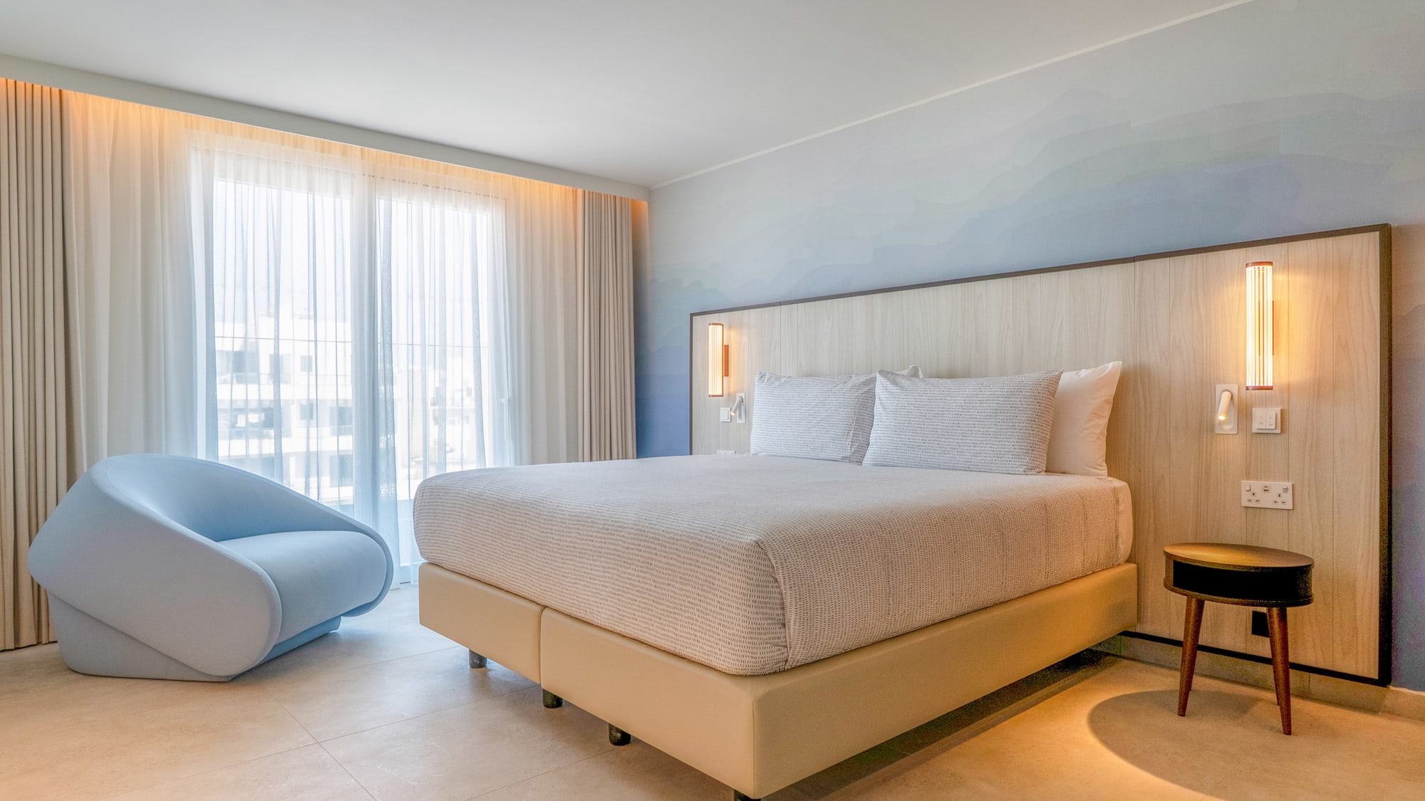 Modern hotel bedroom with king bed, light wood headboard, blue accent chair, large window.