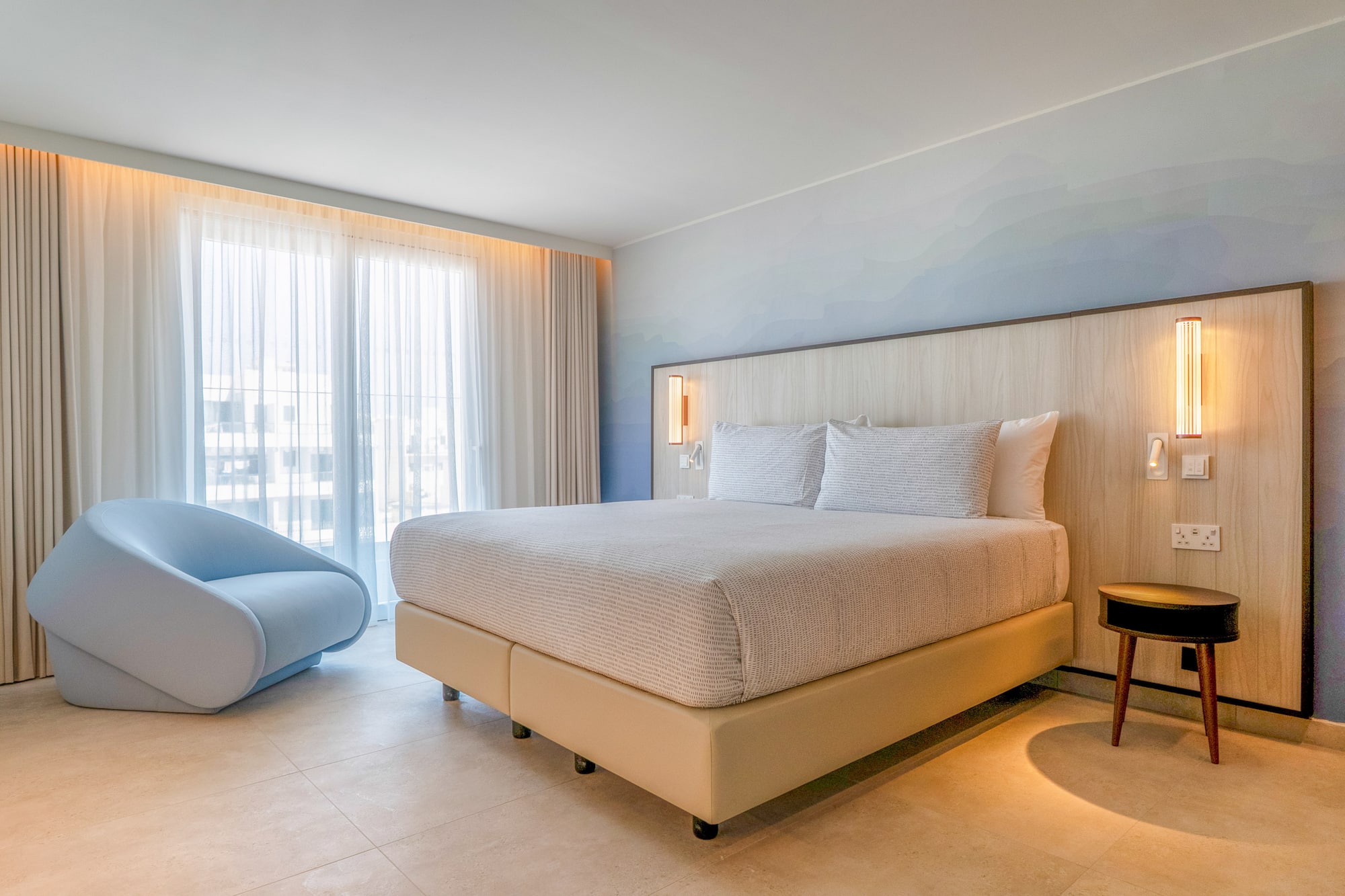 Modern hotel bedroom with king bed, light wood headboard, blue accent chair, large window.
