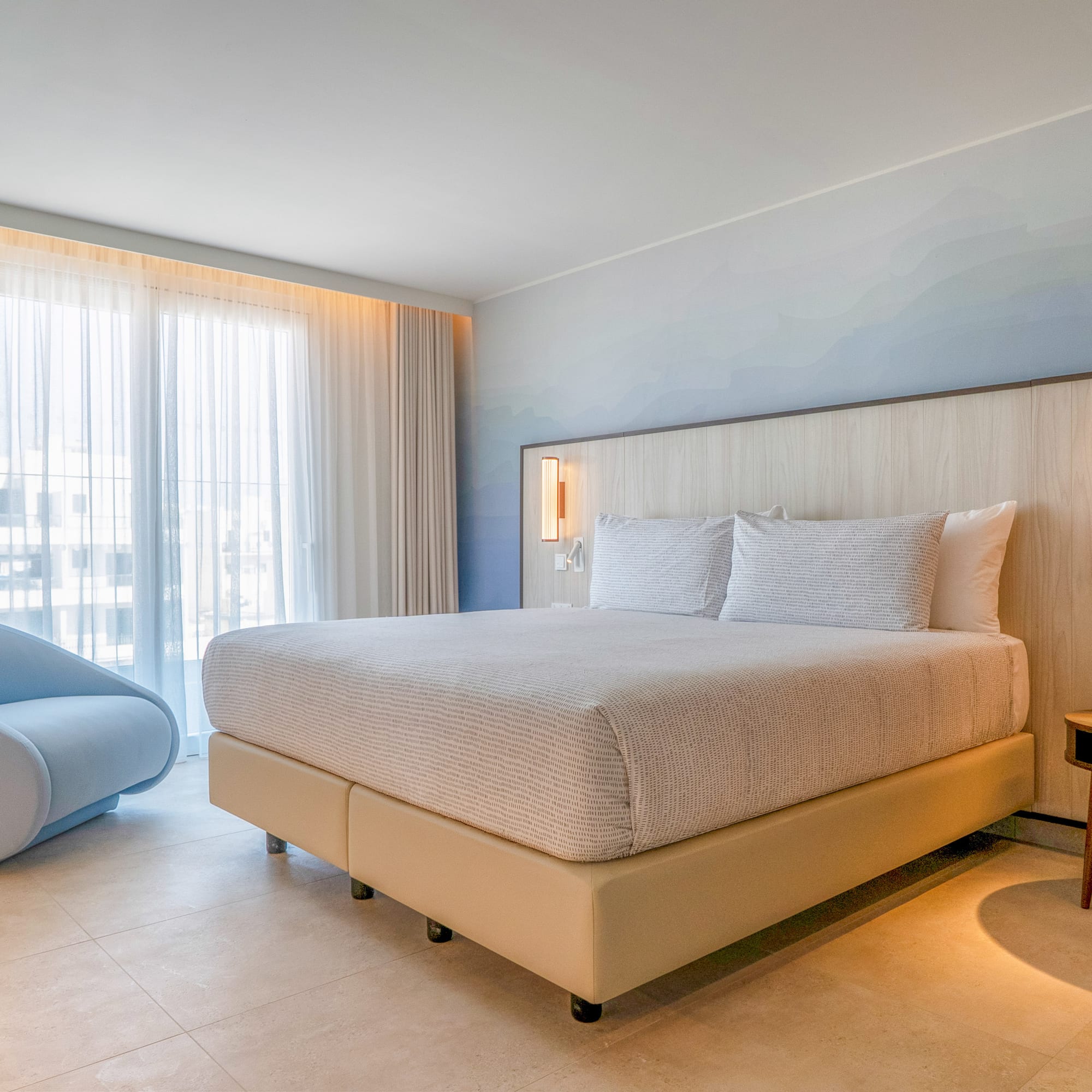 Modern hotel bedroom with king bed, light wood headboard, blue accent chair, large window.