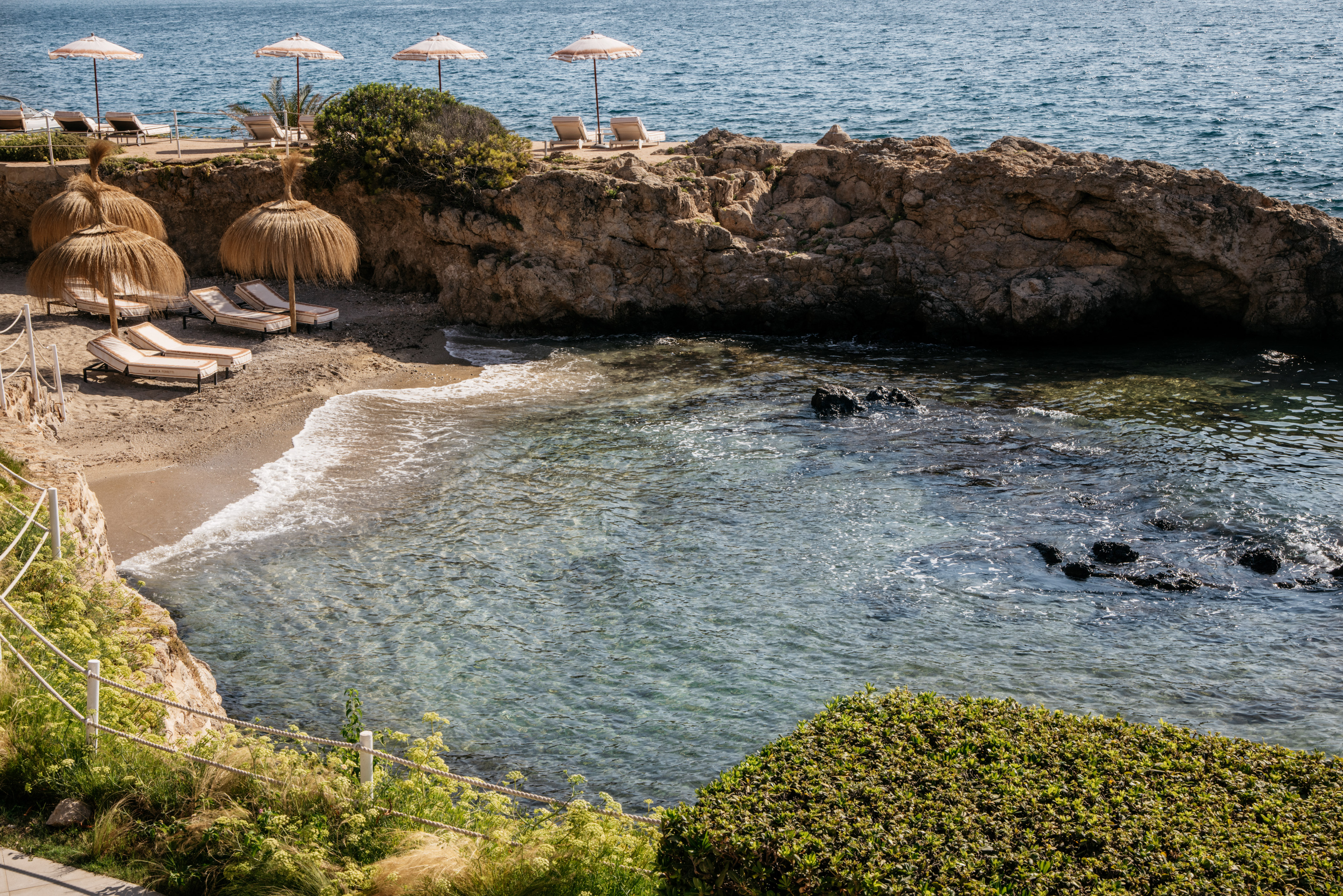 Sandy cove with sun loungers, straw umbrellas, rocky cliffs, and clear sea.