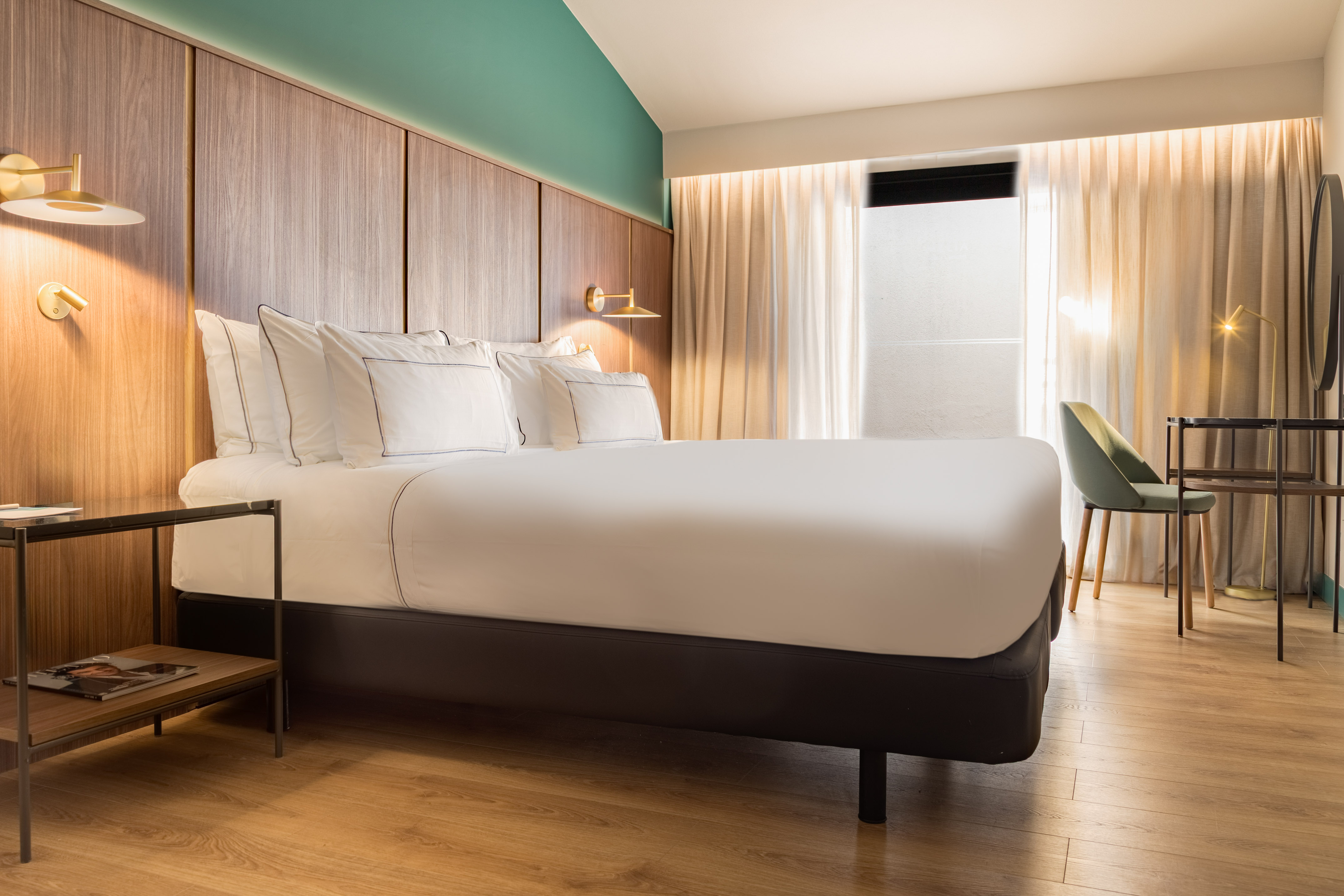 Modern hotel room with king bed, wood headboard, teal wall, and light wood floor.