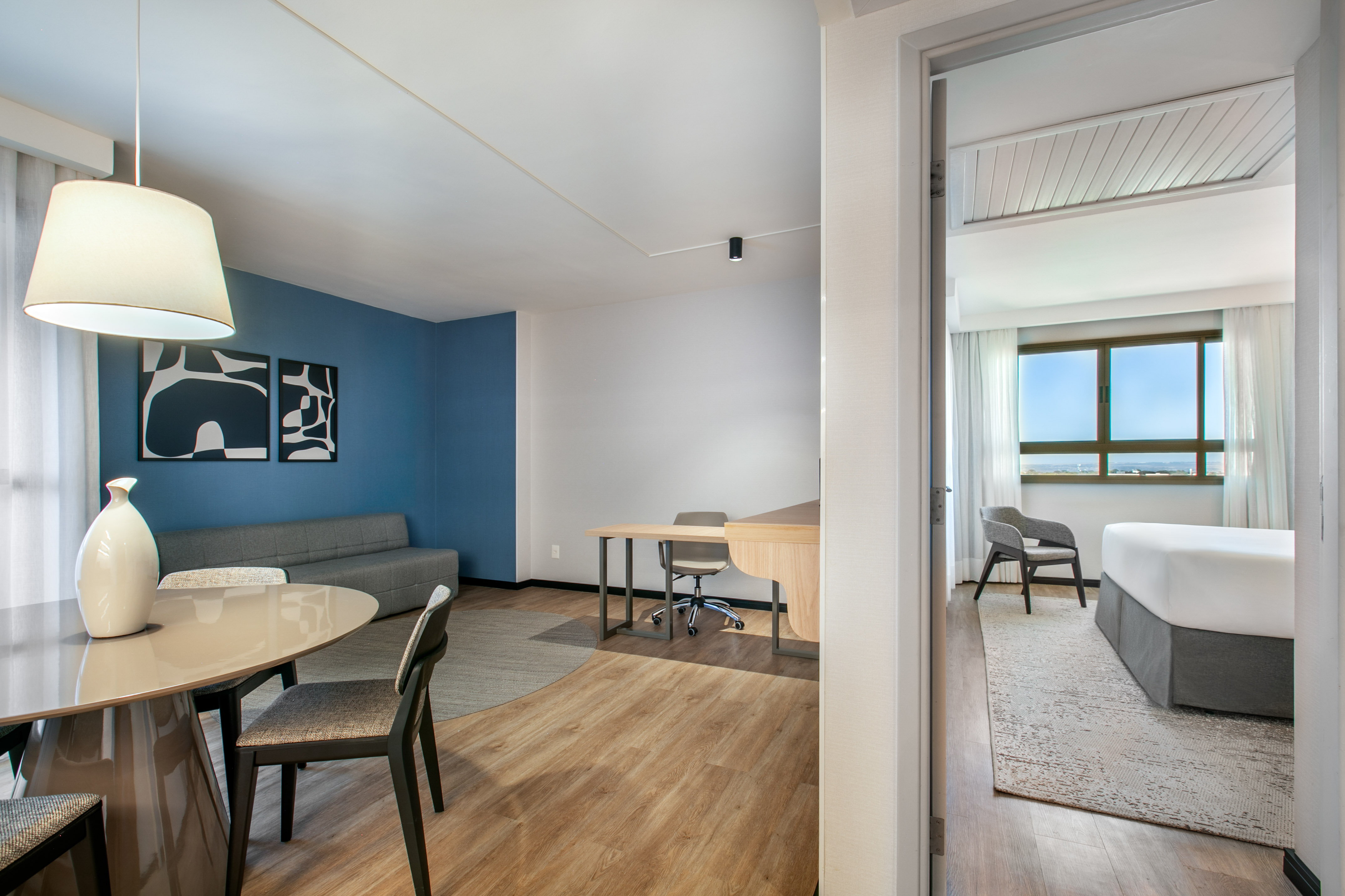 Contemporary hotel suite with living, dining, and workspace, leading to a bedroom.