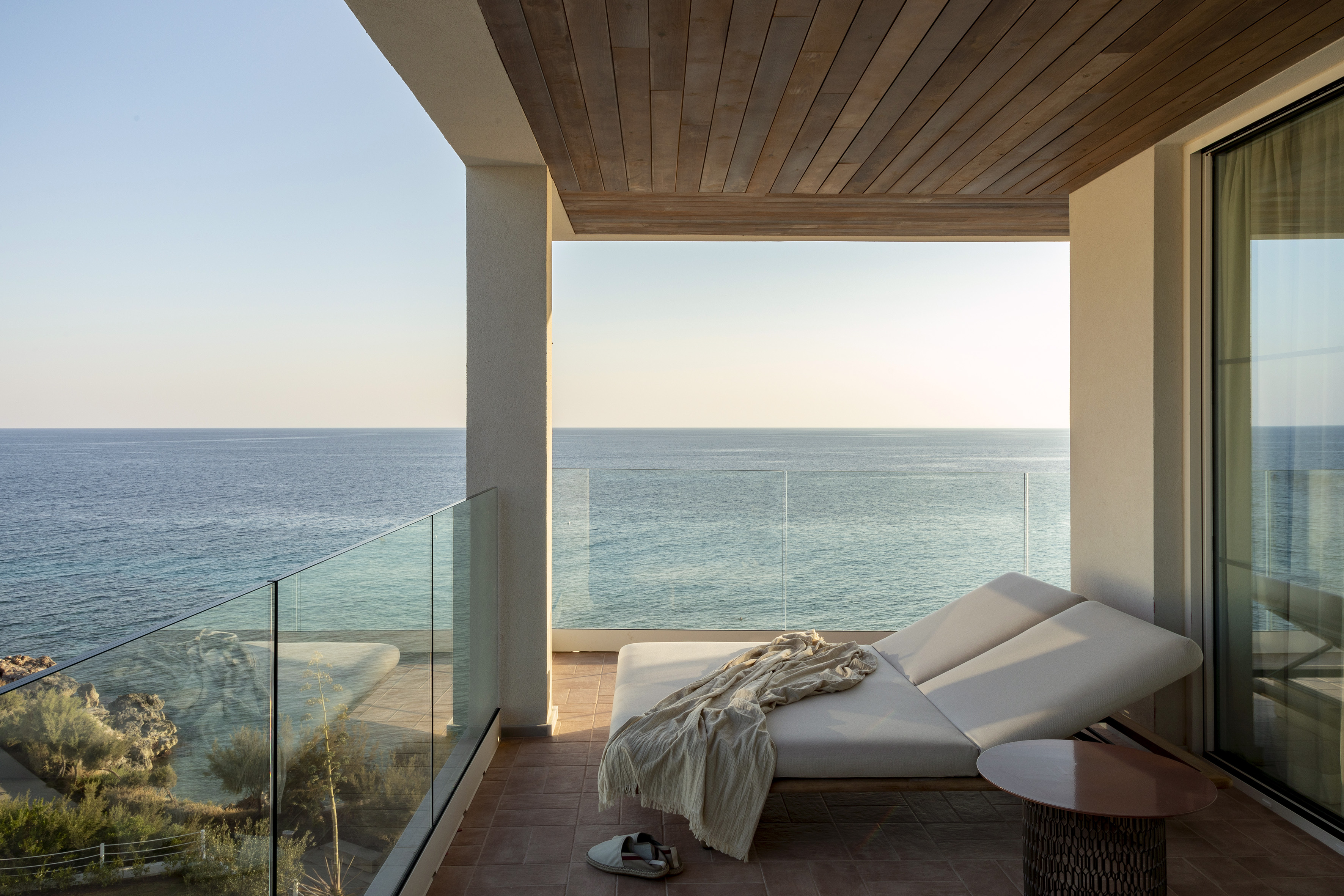 Oceanfront balcony with daybed, glass railing, and clear blue sea.