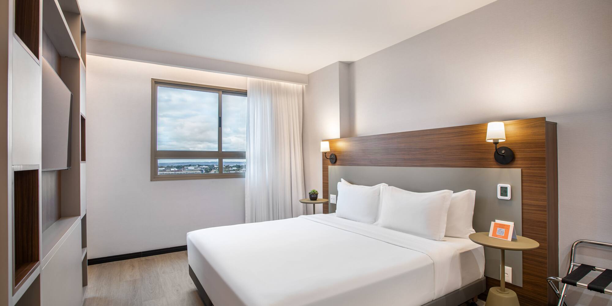 Modern hotel room with queen bed, wood headboard, and city view window.