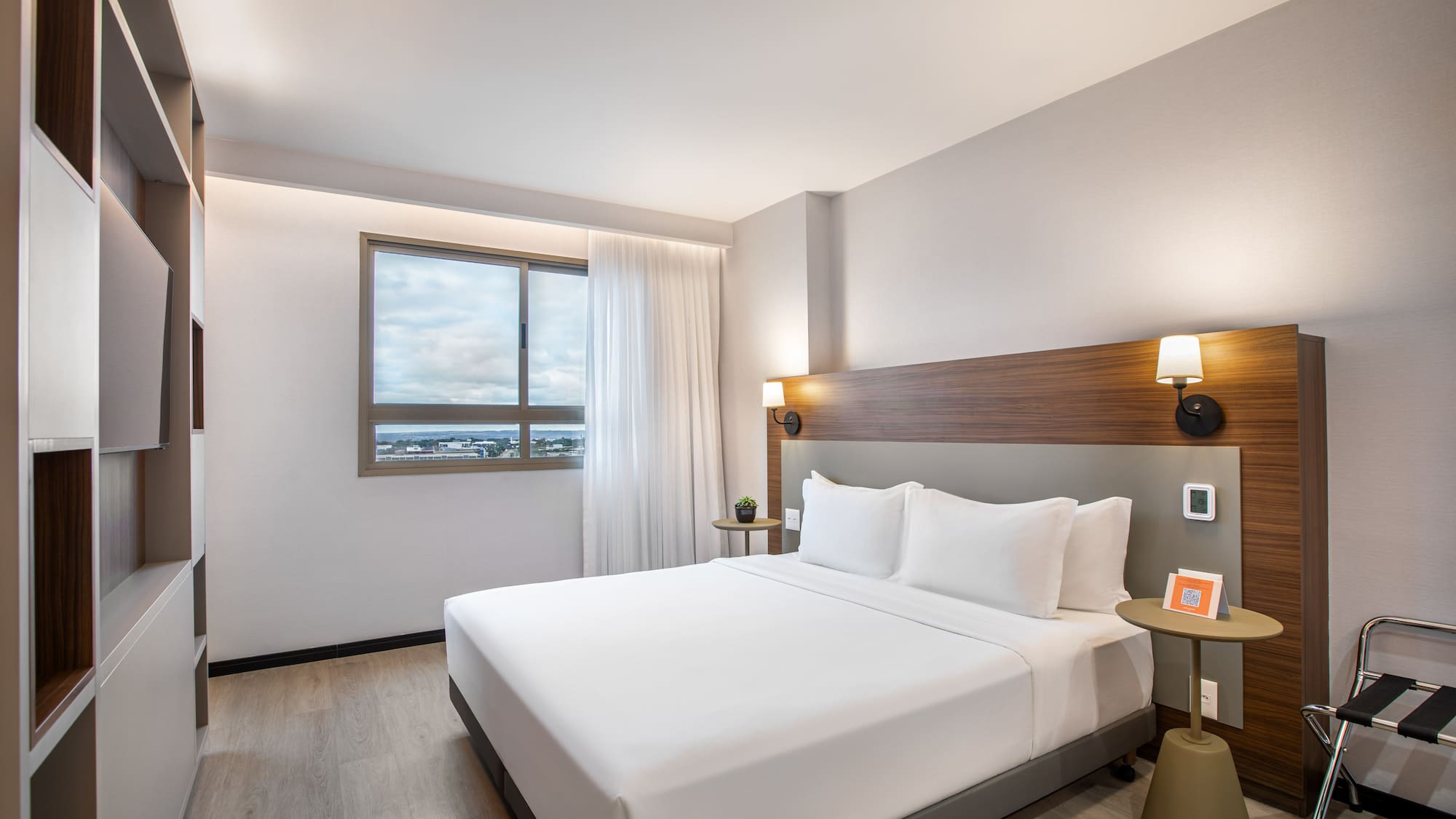 Modern hotel room with queen bed, wood headboard, and city view window.