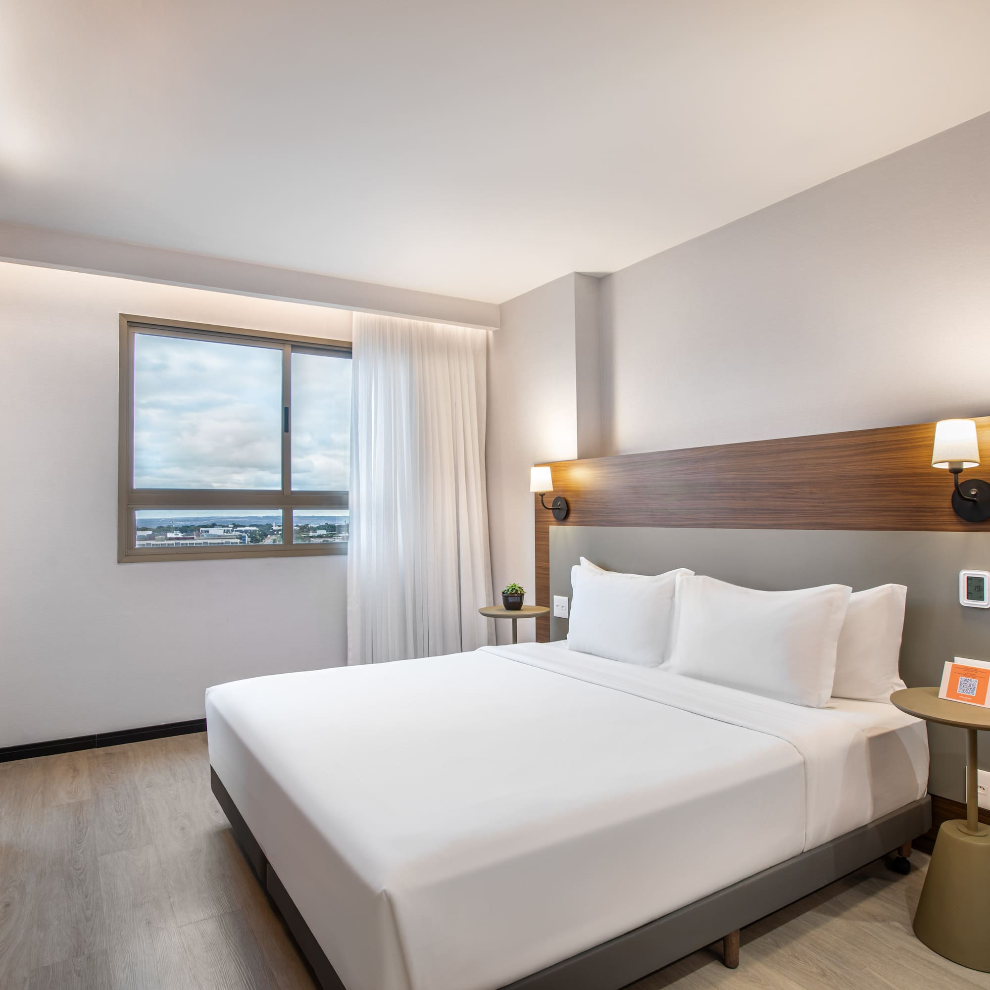 Modern hotel room with queen bed, wood headboard, and city view window.