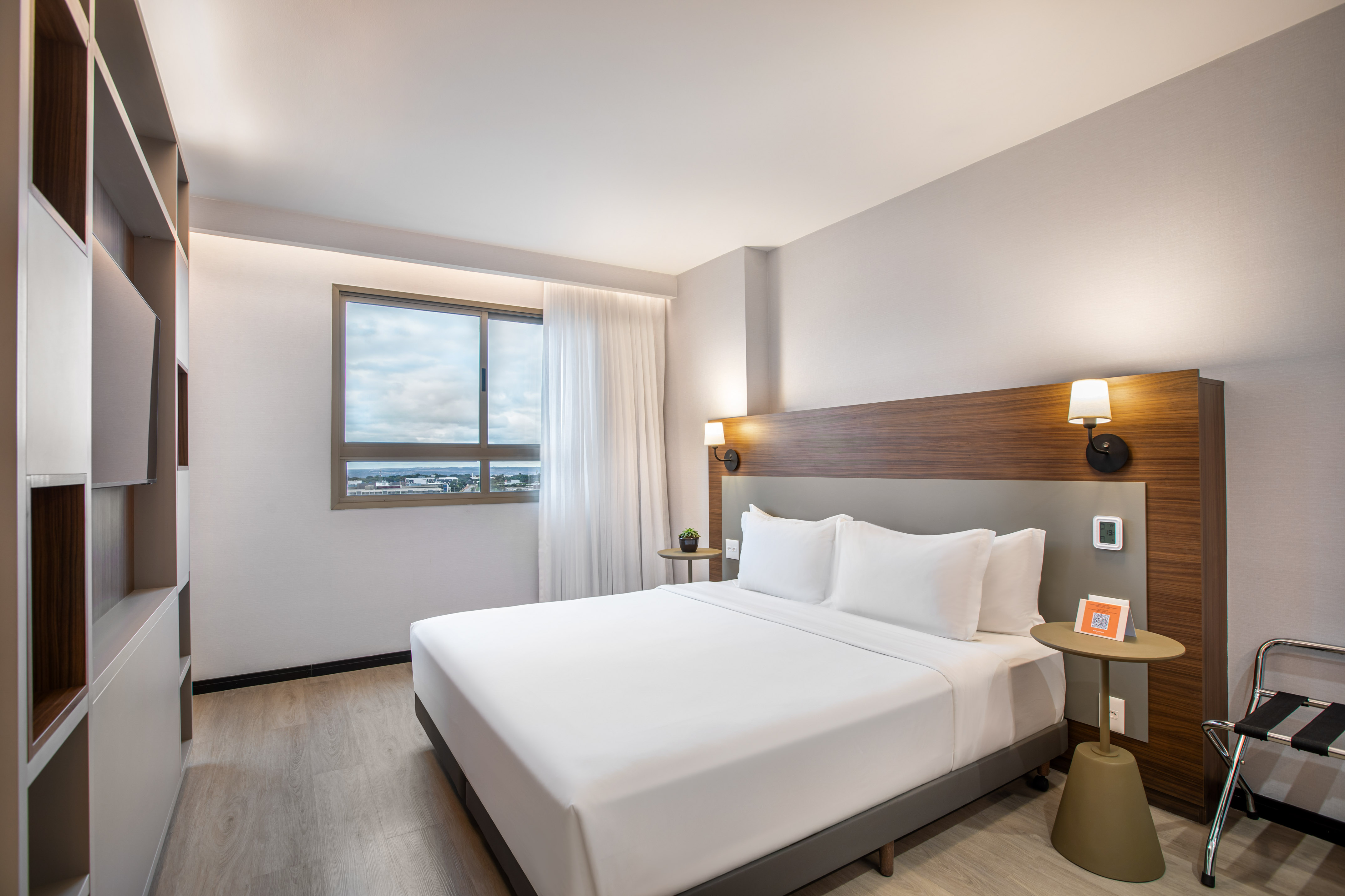Modern hotel room with queen bed, wood headboard, and city view window.