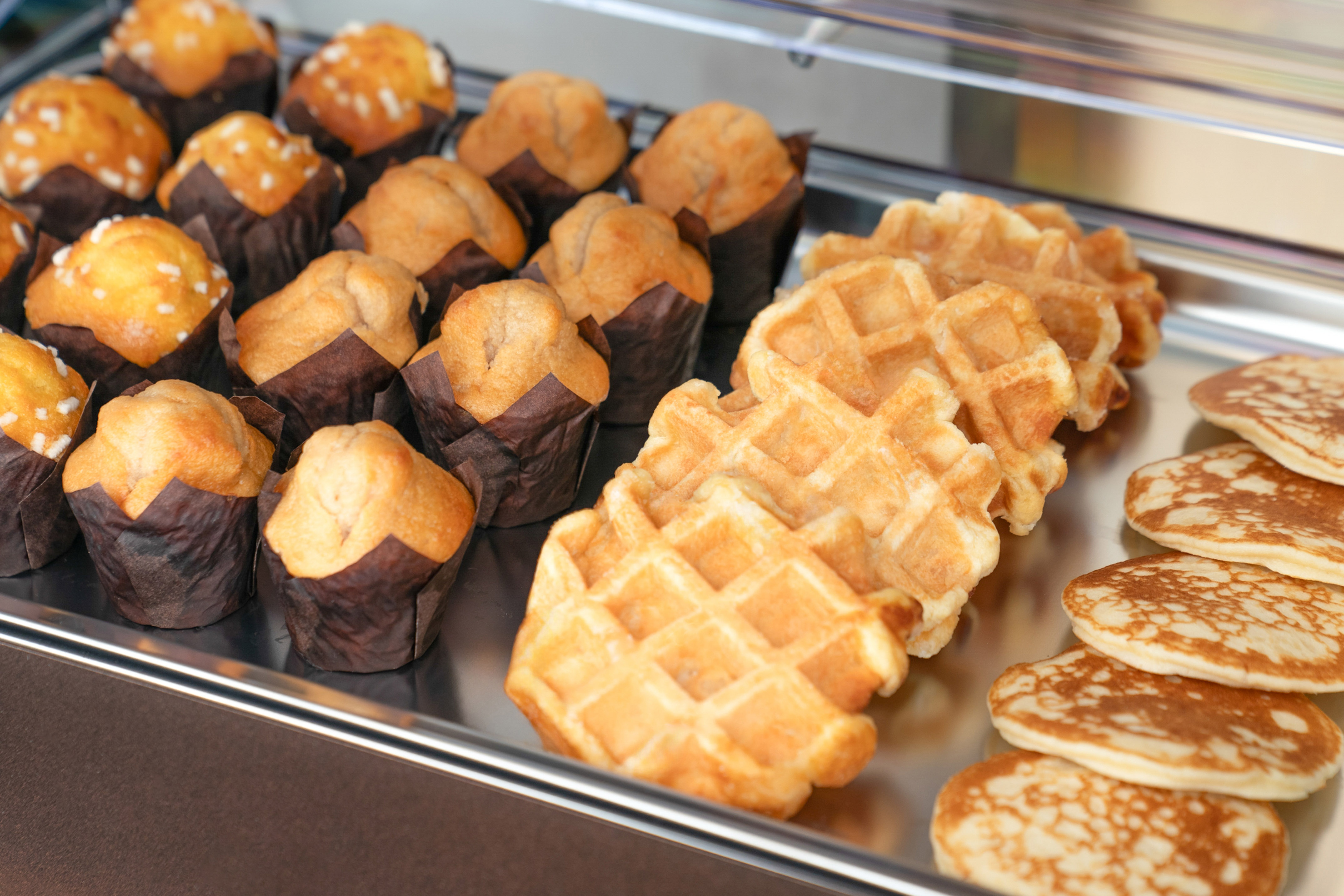 Warm muffins, golden waffles, and small pancakes on a breakfast buffet tray.