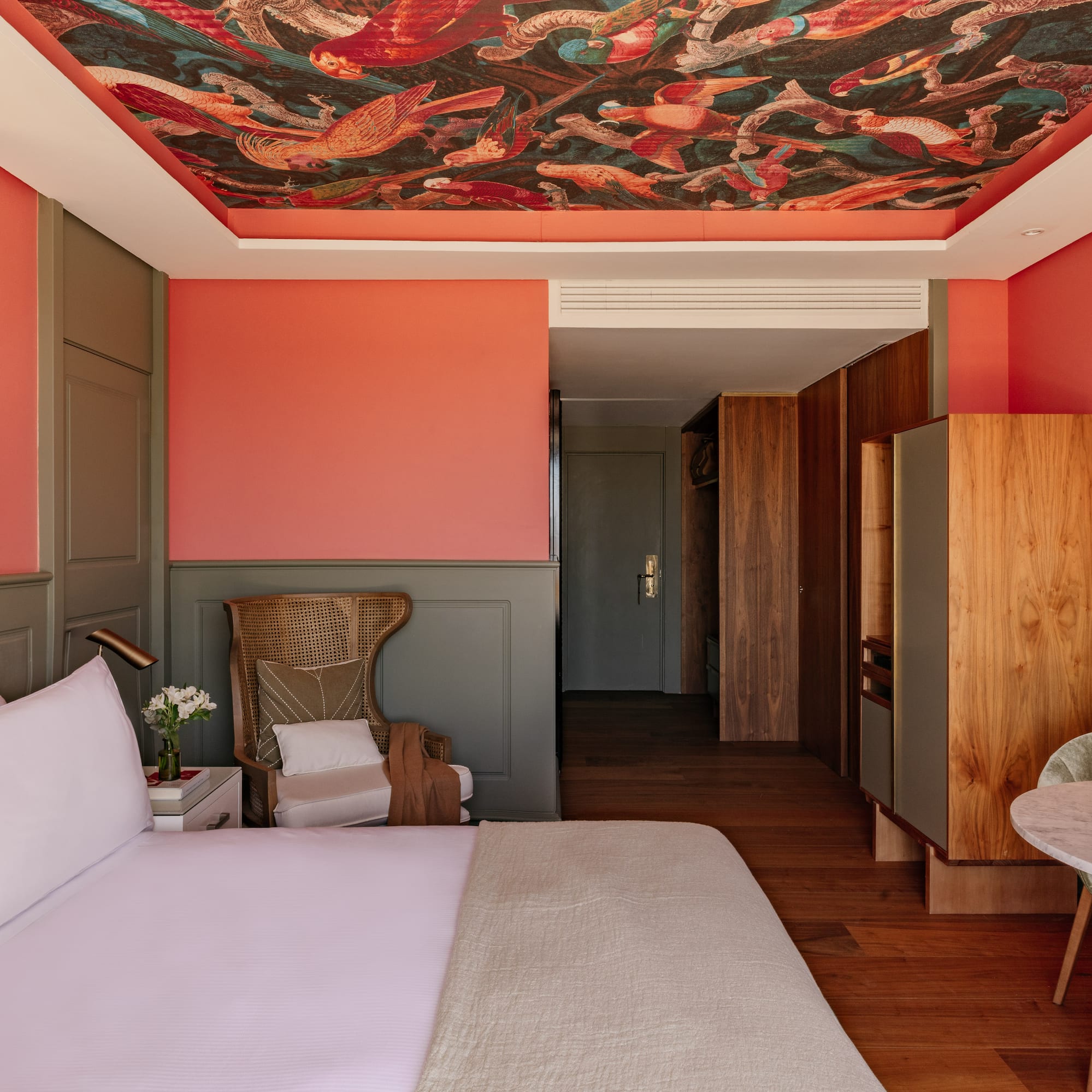Dormitorio hotel con mural de techo, paredes coral, cabecero y mobiliario.