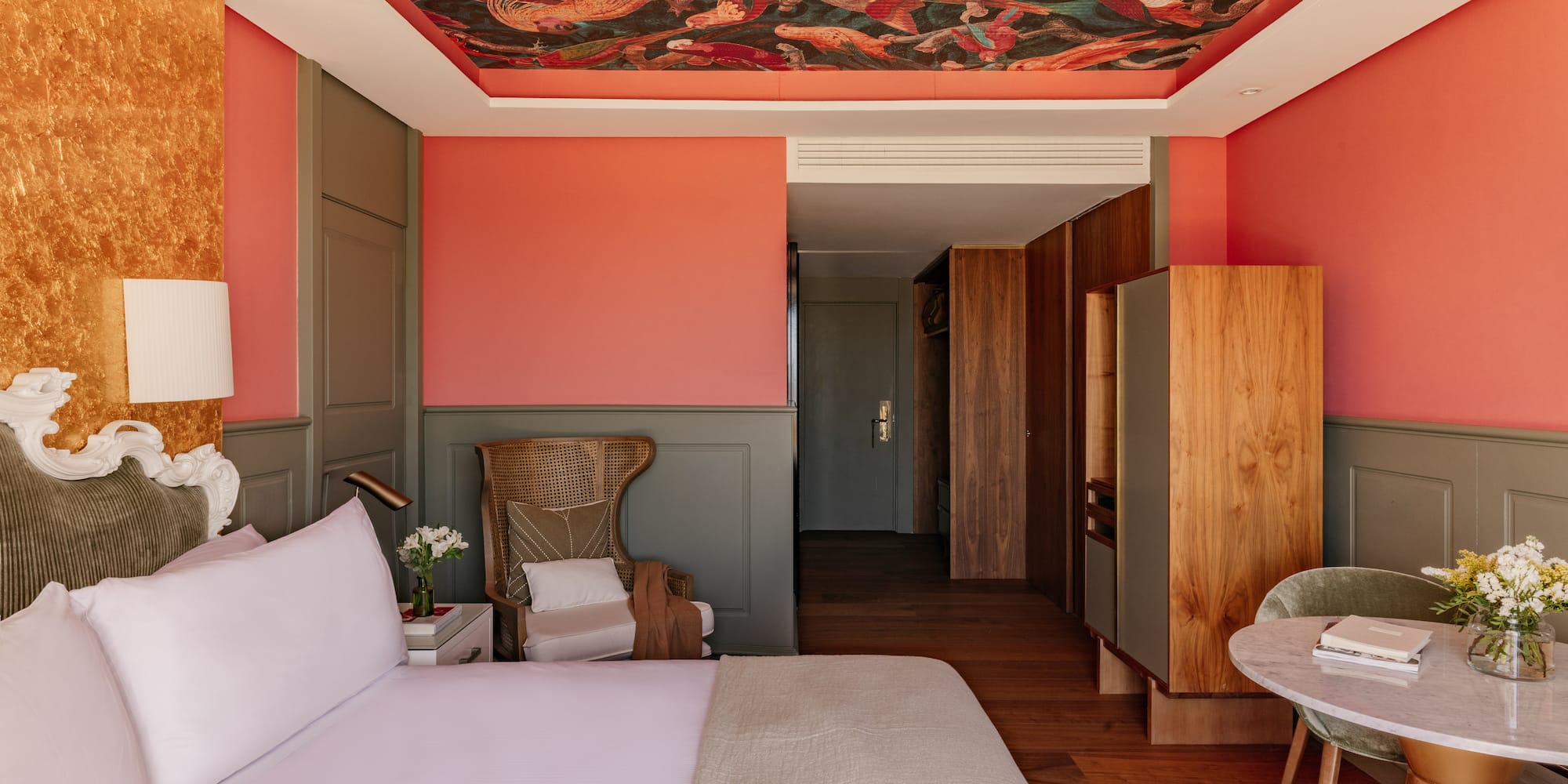 Dormitorio hotel con mural de techo, paredes coral, cabecero y mobiliario.