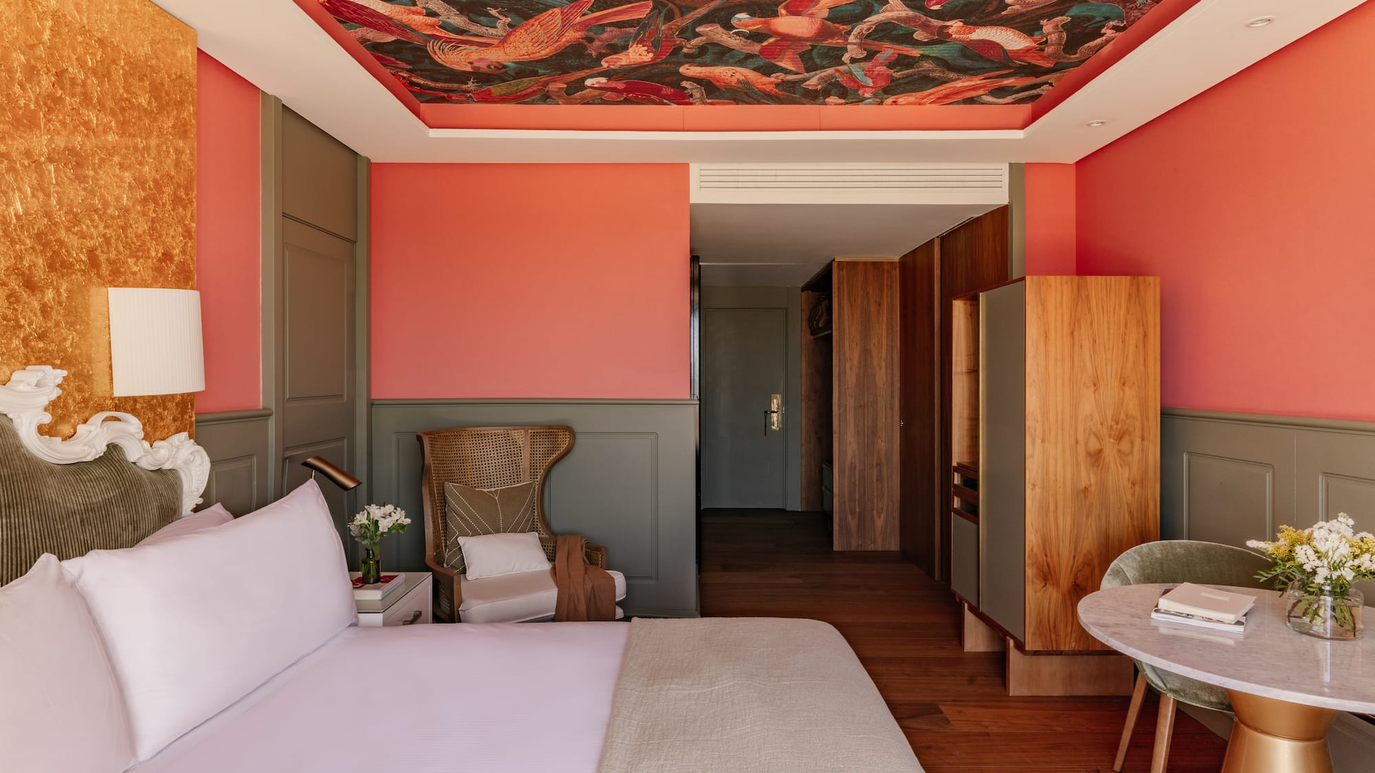 Dormitorio hotel con mural de techo, paredes coral, cabecero y mobiliario.
