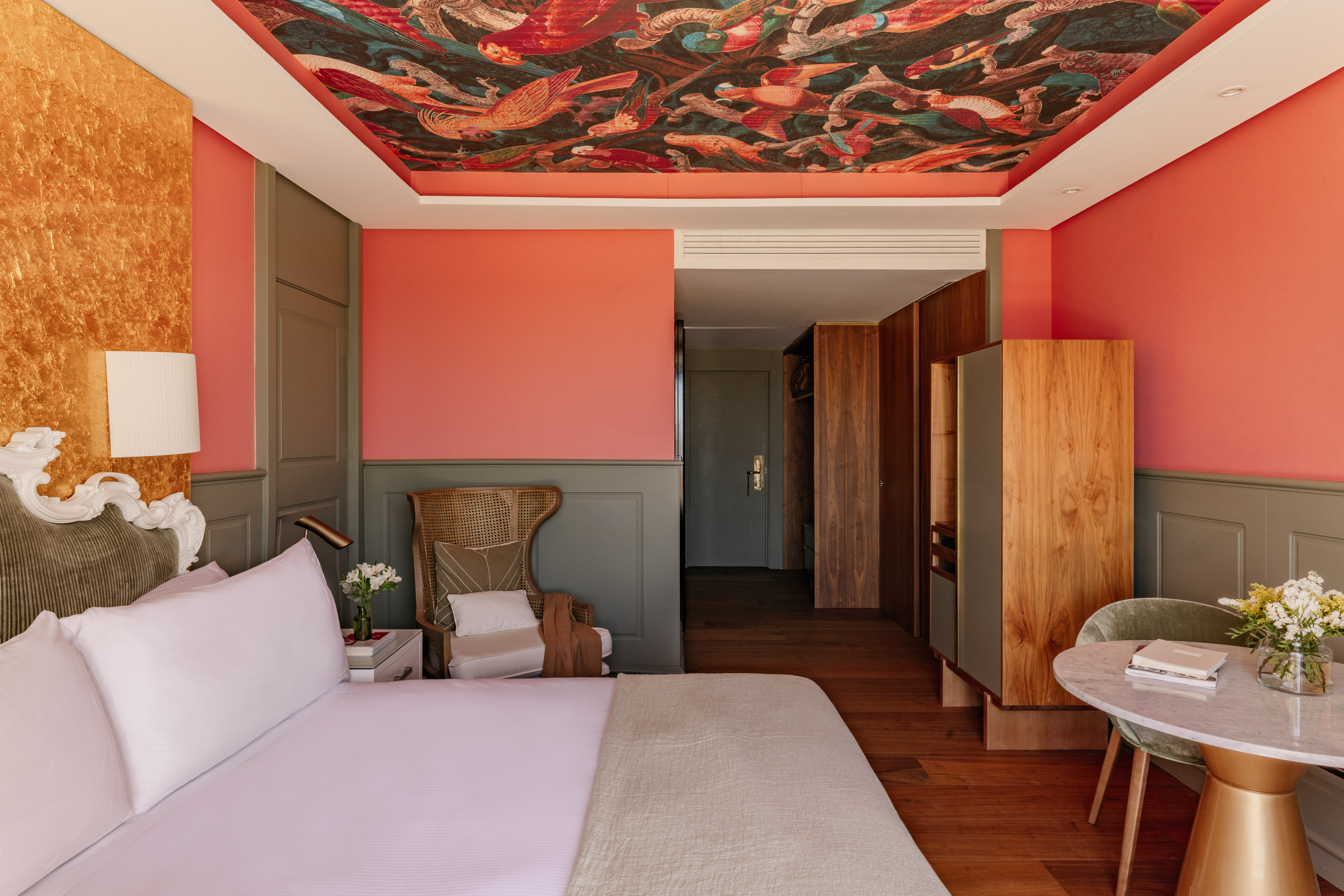 Dormitorio hotel con mural de techo, paredes coral, cabecero y mobiliario.