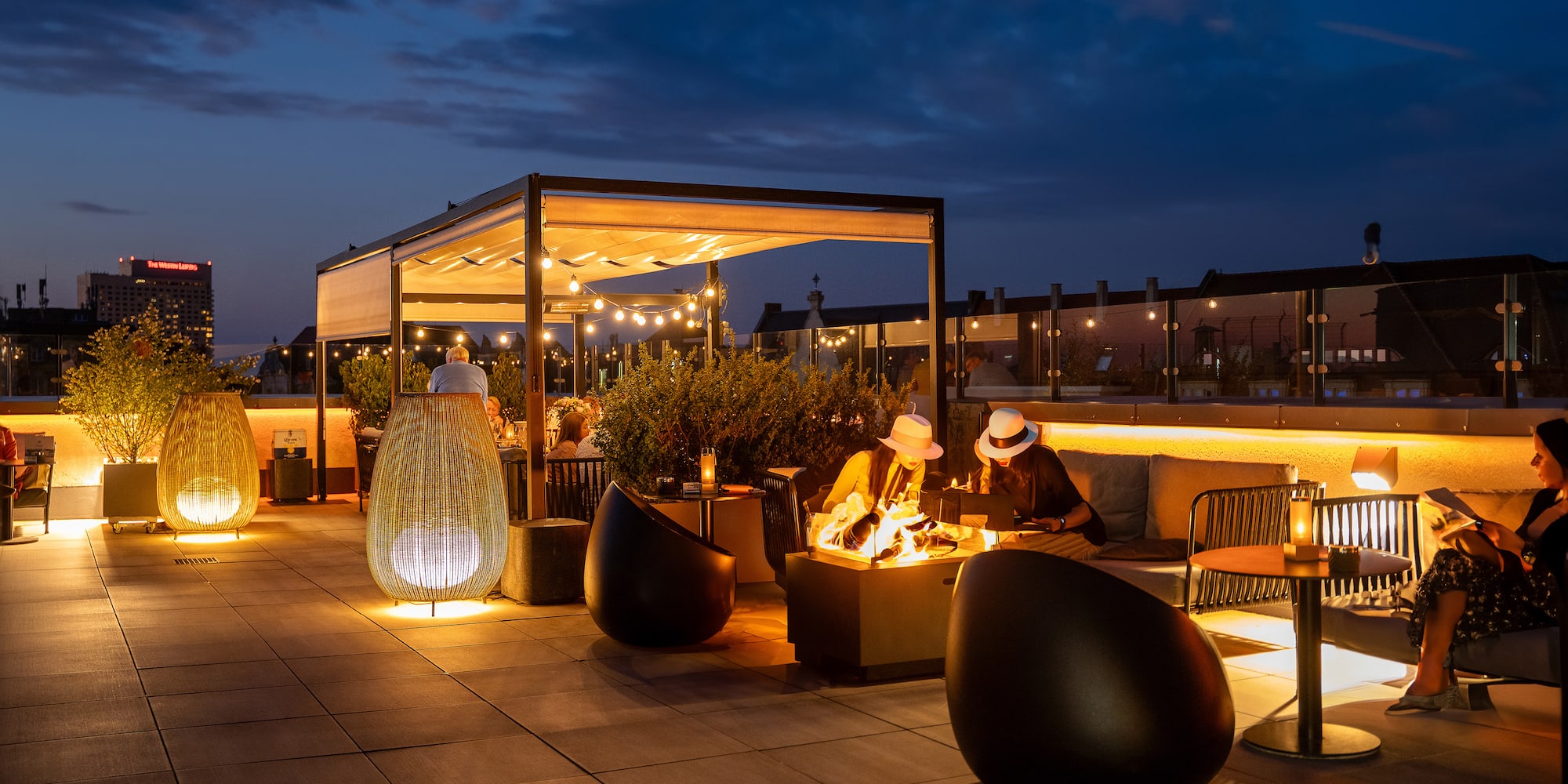Rooftop bar at dusk with fire pit, string lights, cozy seating, and city skyline.