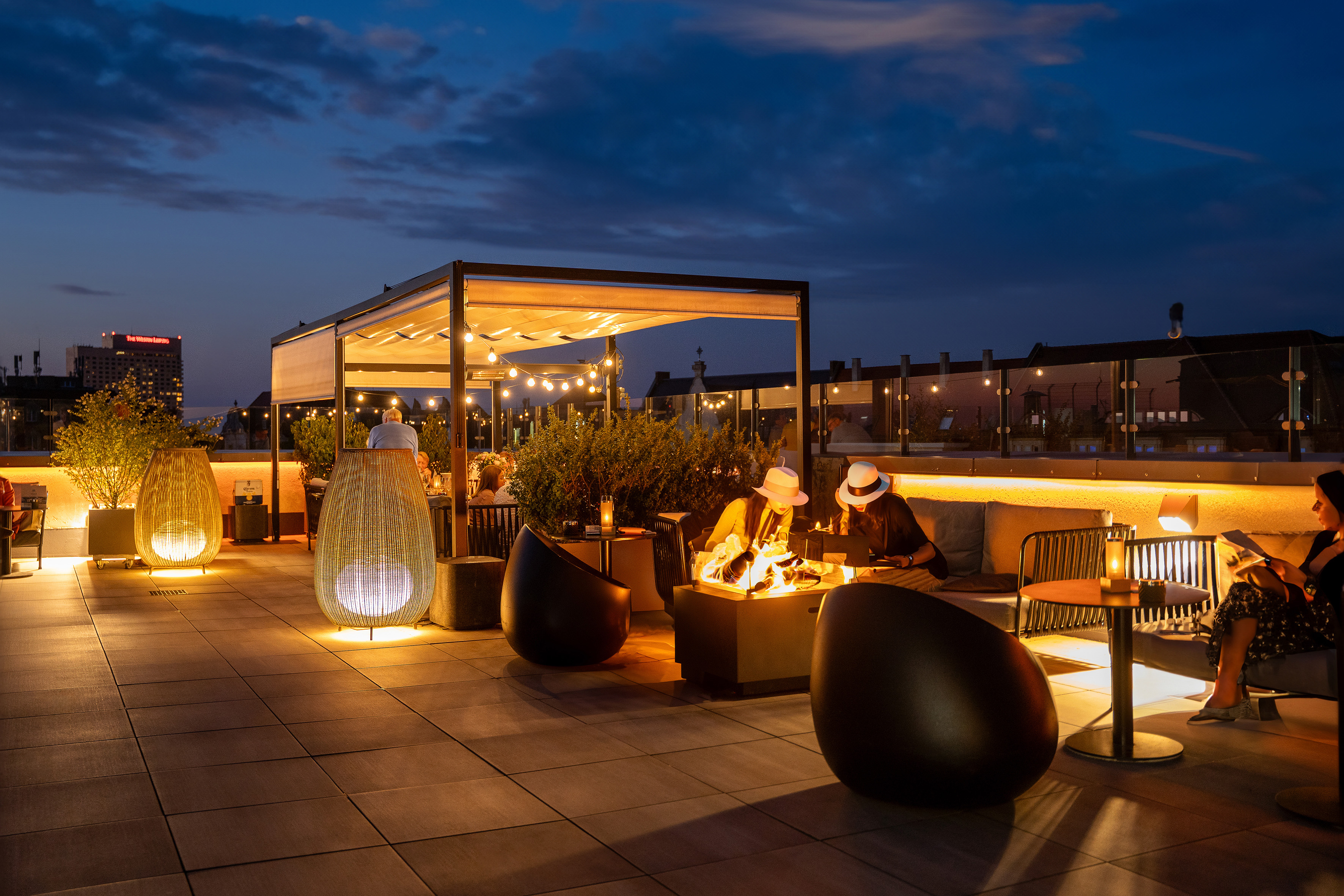 Rooftop bar at dusk with fire pit, string lights, cozy seating, and city skyline.