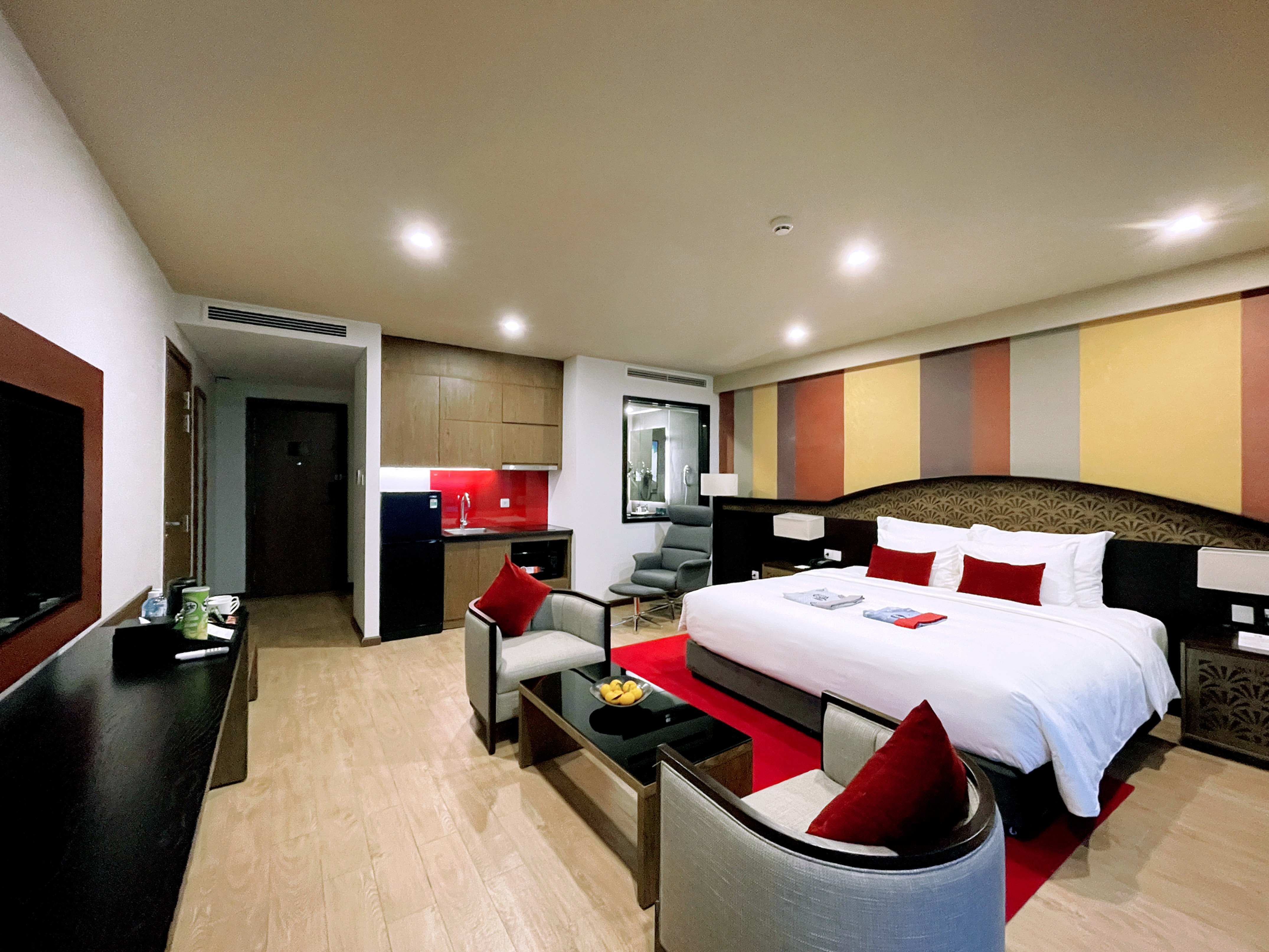 Modern hotel room with king bed, red rug, cozy sitting area, and kitchenette.