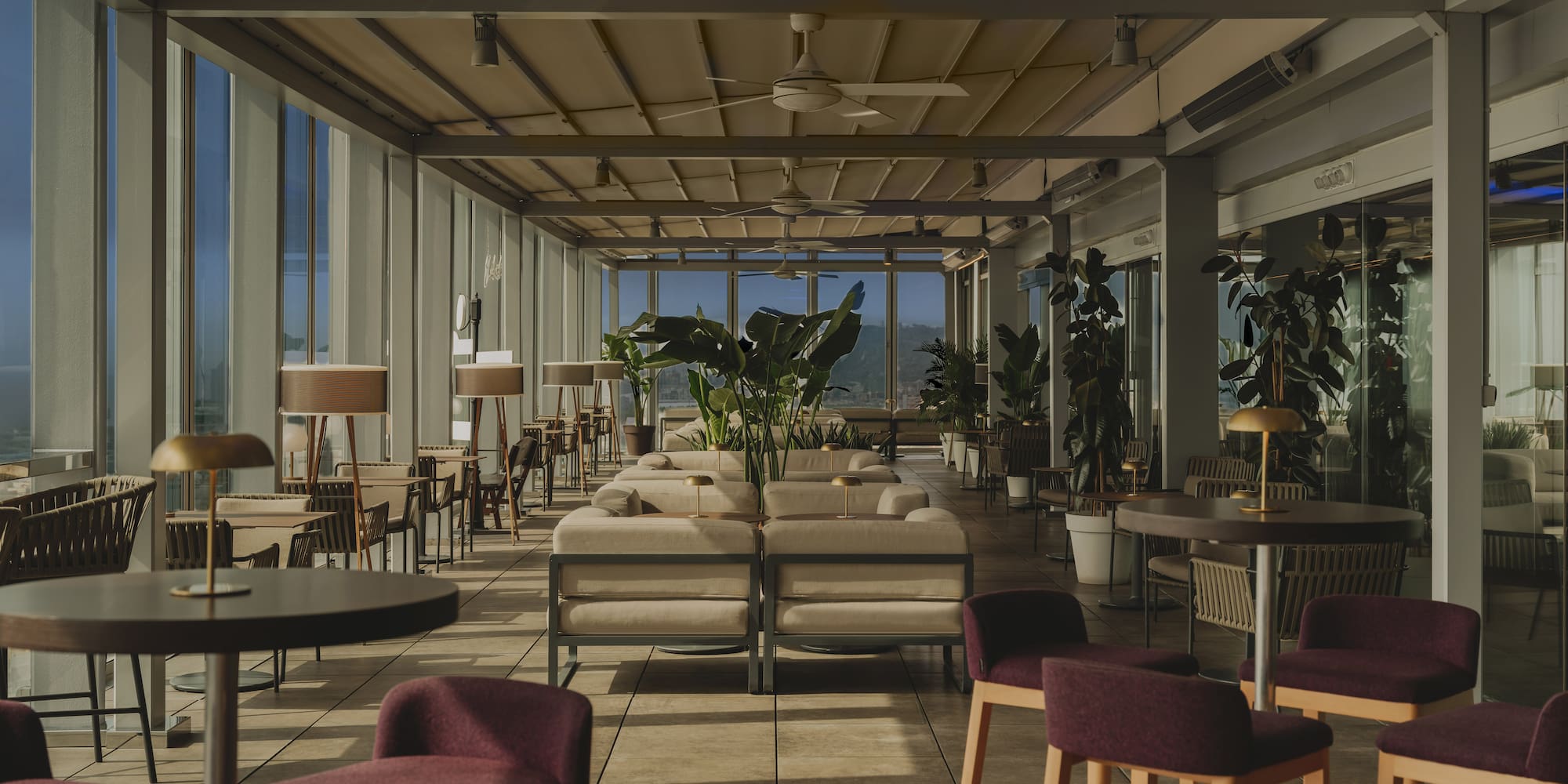 Rooftop bar lounge with stylish seating, ceiling fans, plants, and bright window views.