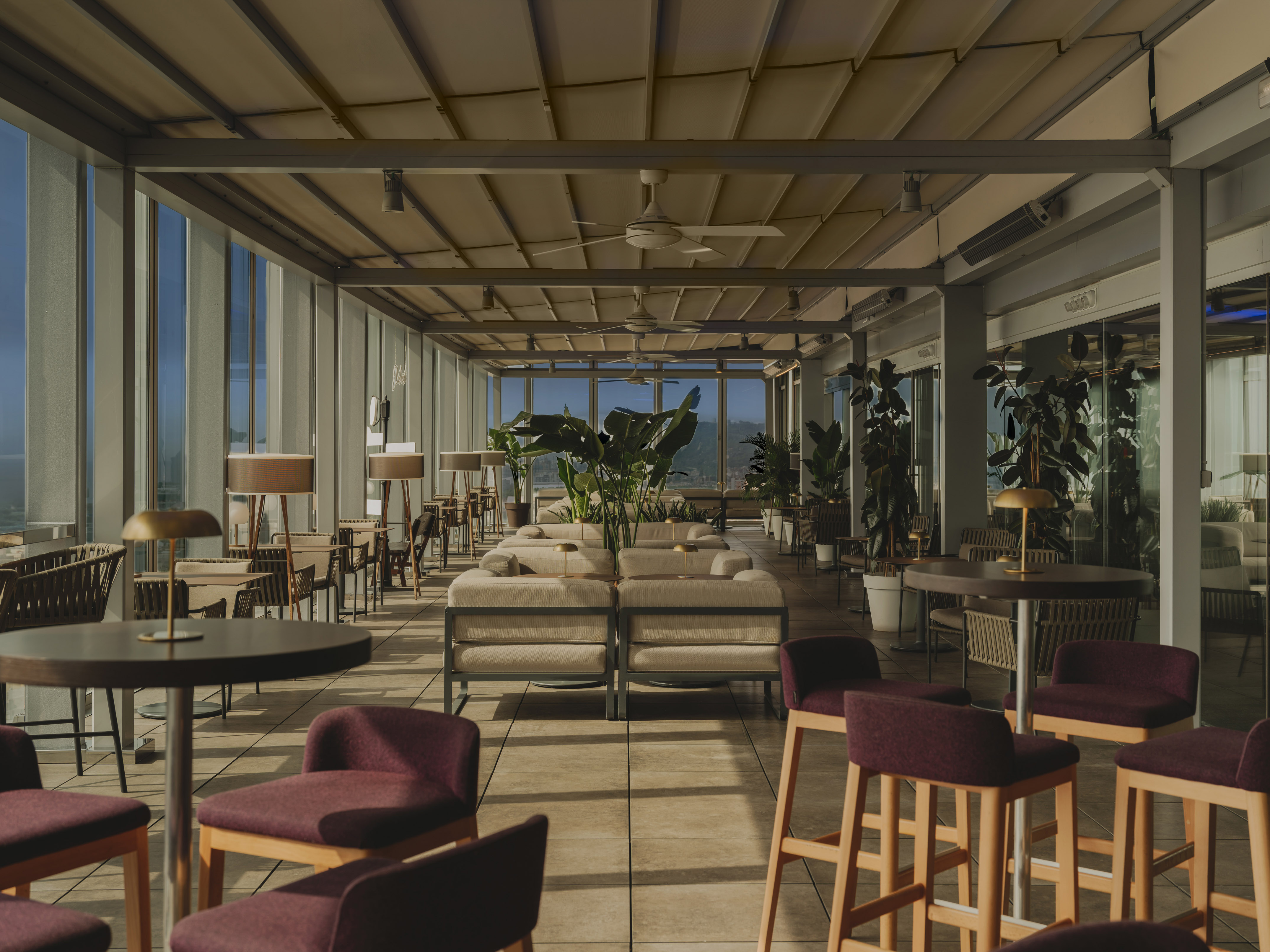Rooftop bar lounge with stylish seating, ceiling fans, plants, and bright window views.