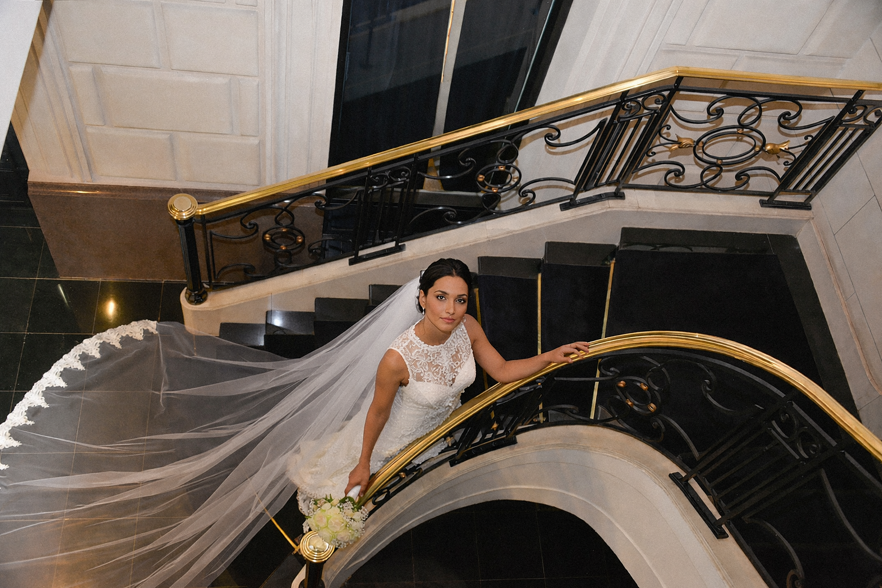 Bride in white lace wedding dress, long veil, on ornate black staircase.