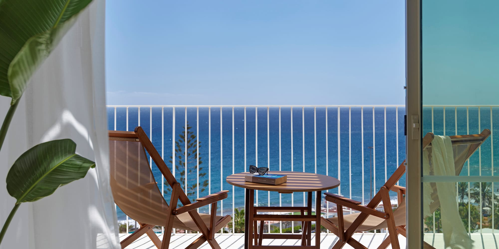 Hotel balcony with sea view, wooden table, chairs, book, and sunglasses.