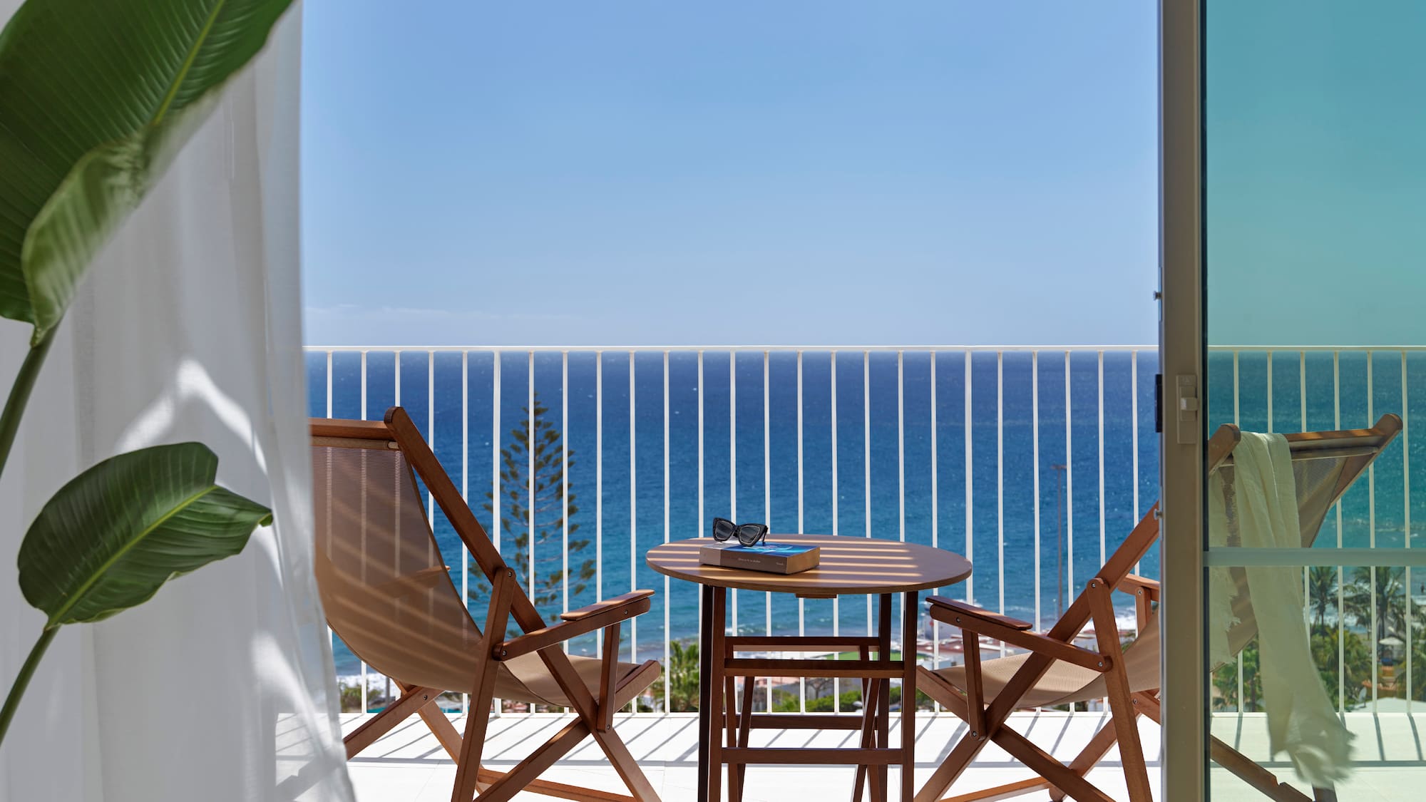 Hotel balcony with sea view, wooden table, chairs, book, and sunglasses.