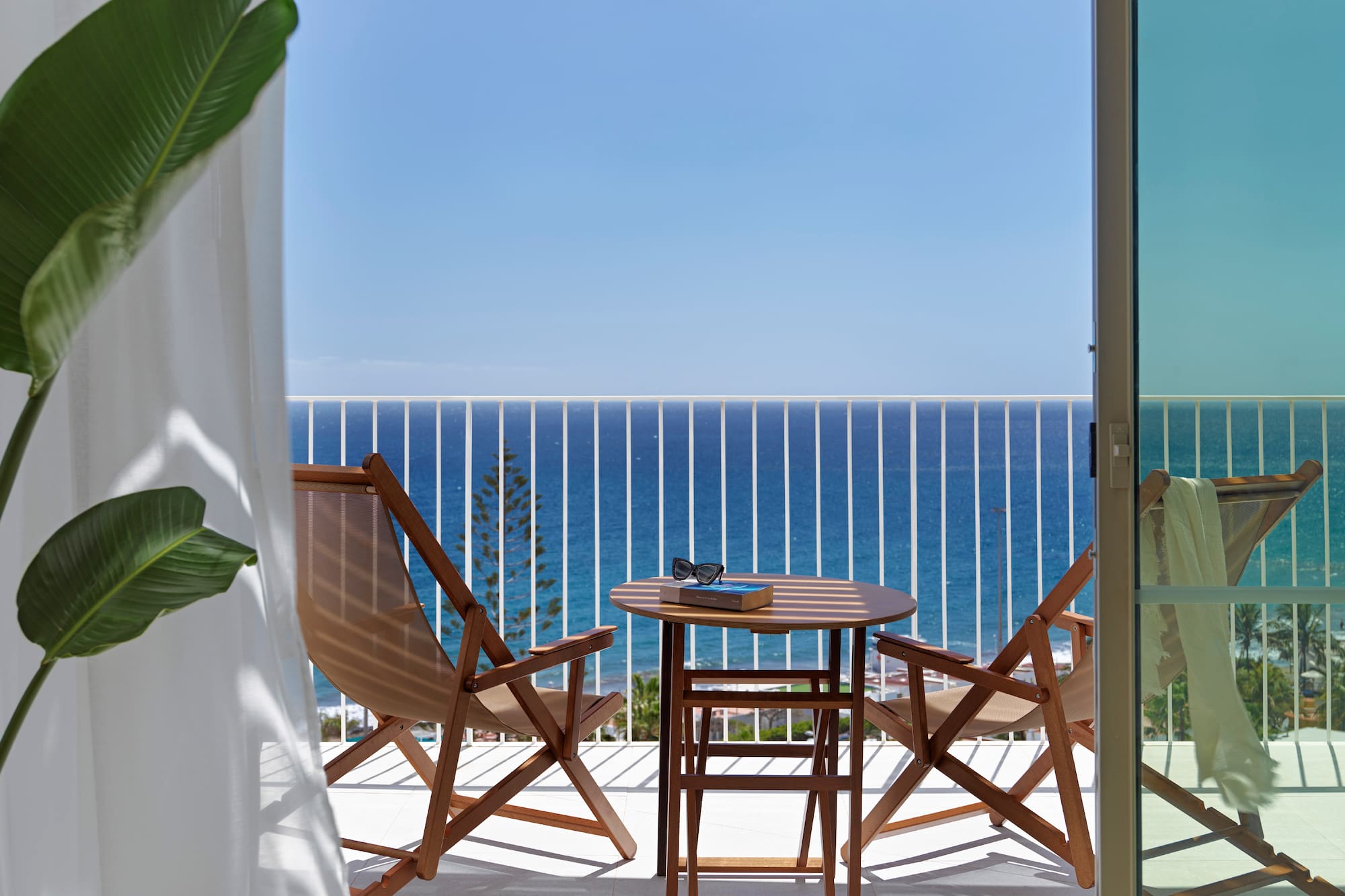 Hotel balcony with sea view, wooden table, chairs, book, and sunglasses.
