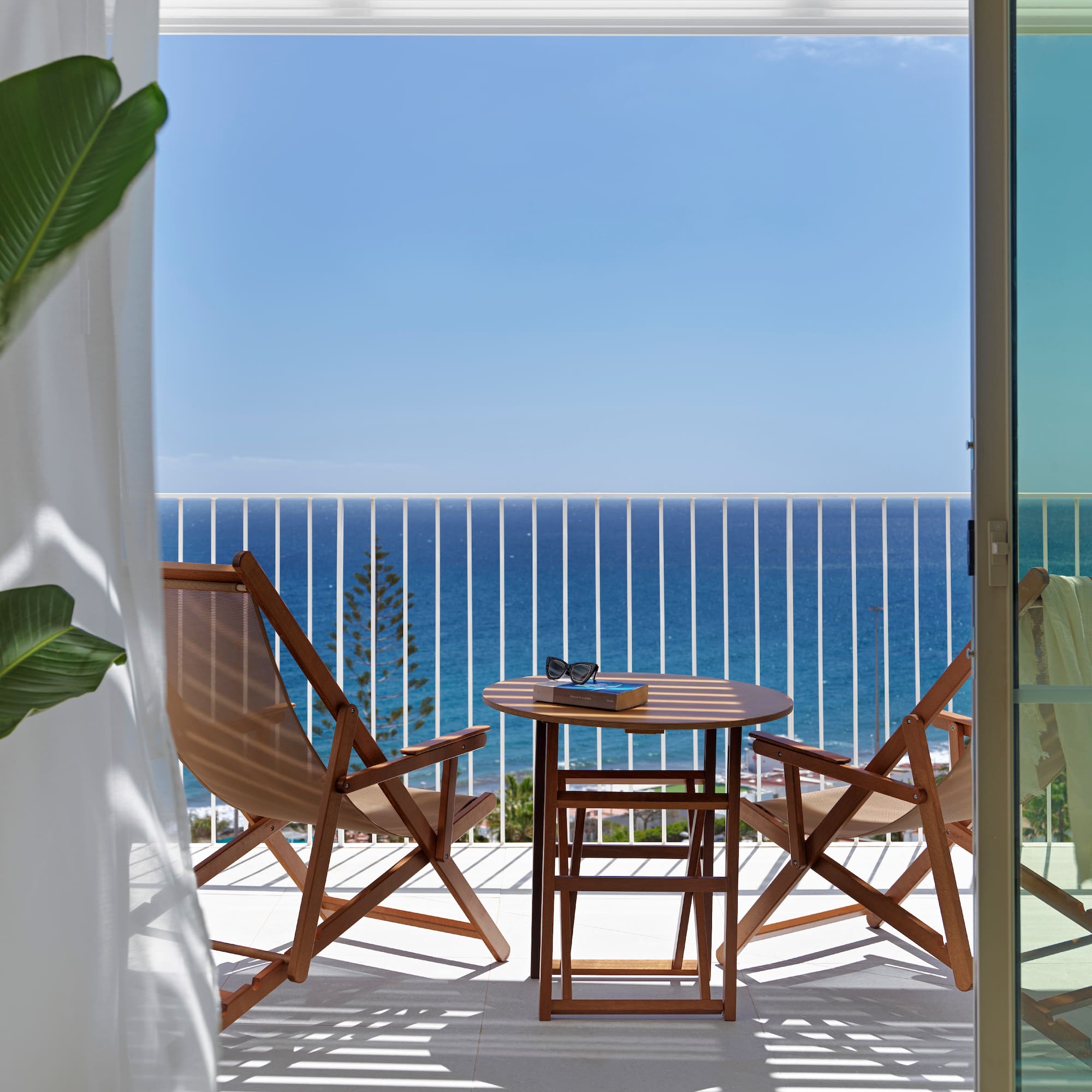 Hotel balcony with sea view, wooden table, chairs, book, and sunglasses.