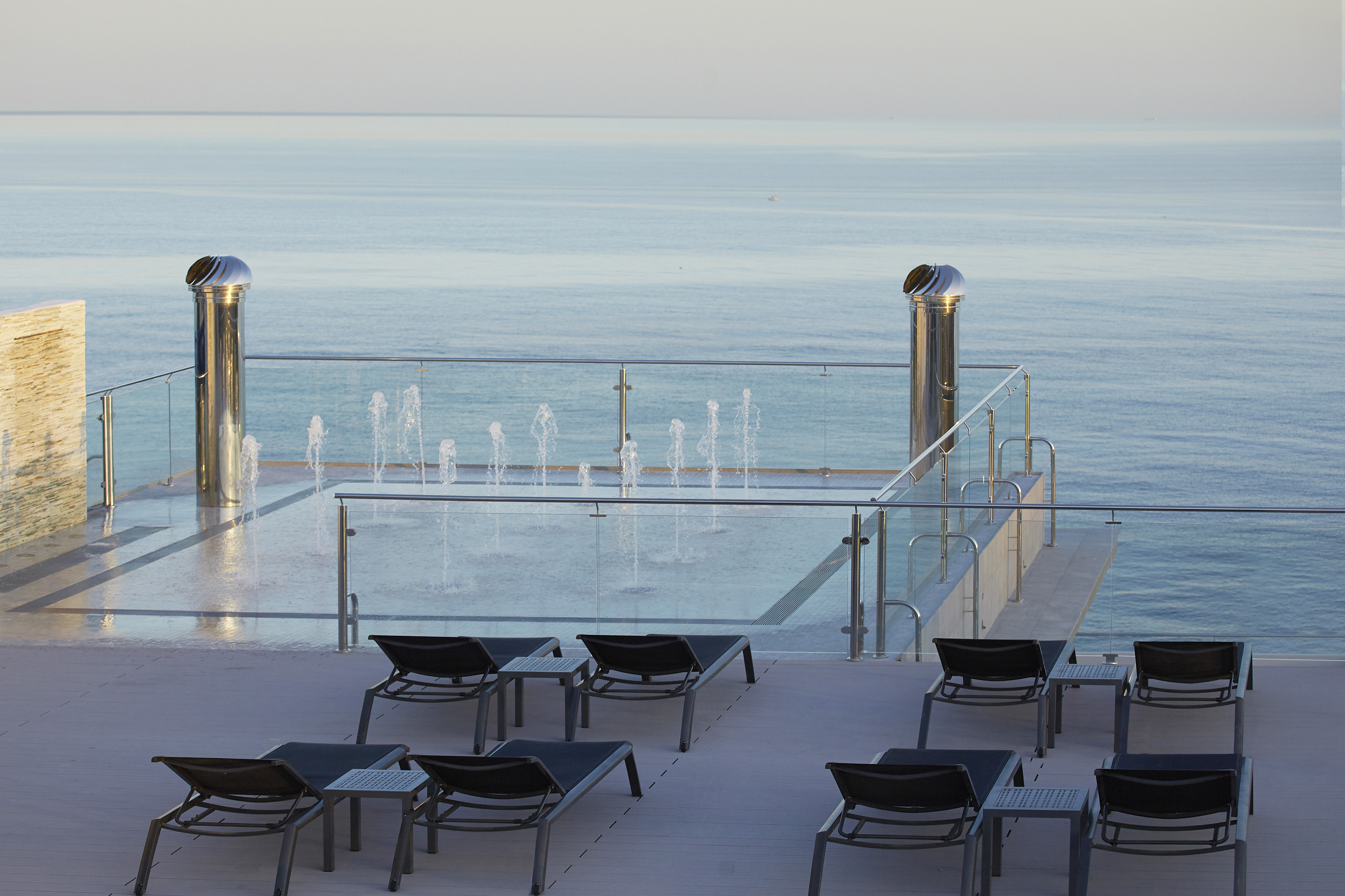 Luxury ocean view terrace with sun loungers, fountains, and glass railing.