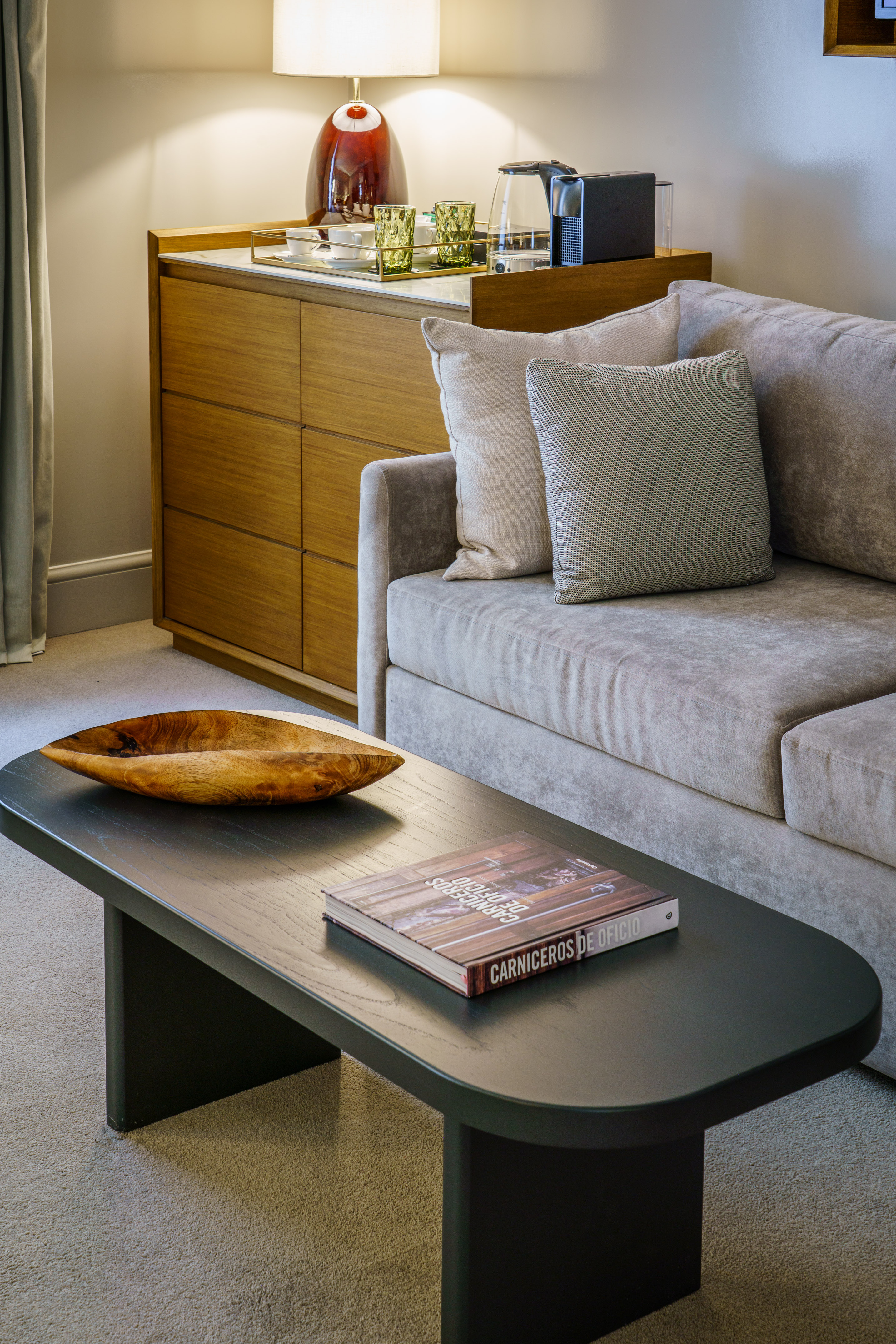 Modern hotel living room with grey sofa, dark coffee table, and refreshment station.