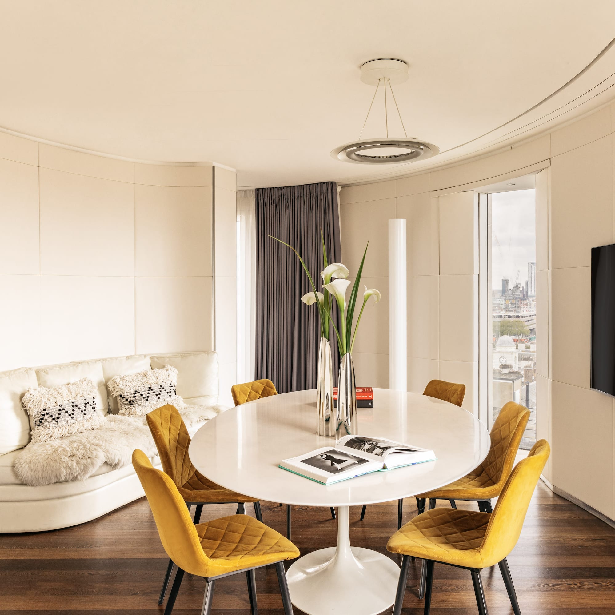 Modern living/dining room with yellow chairs, white table, curved sofa, city view.