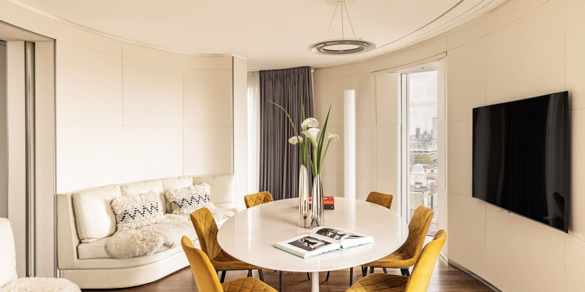 Modern living/dining room with yellow chairs, white table, curved sofa, city view.