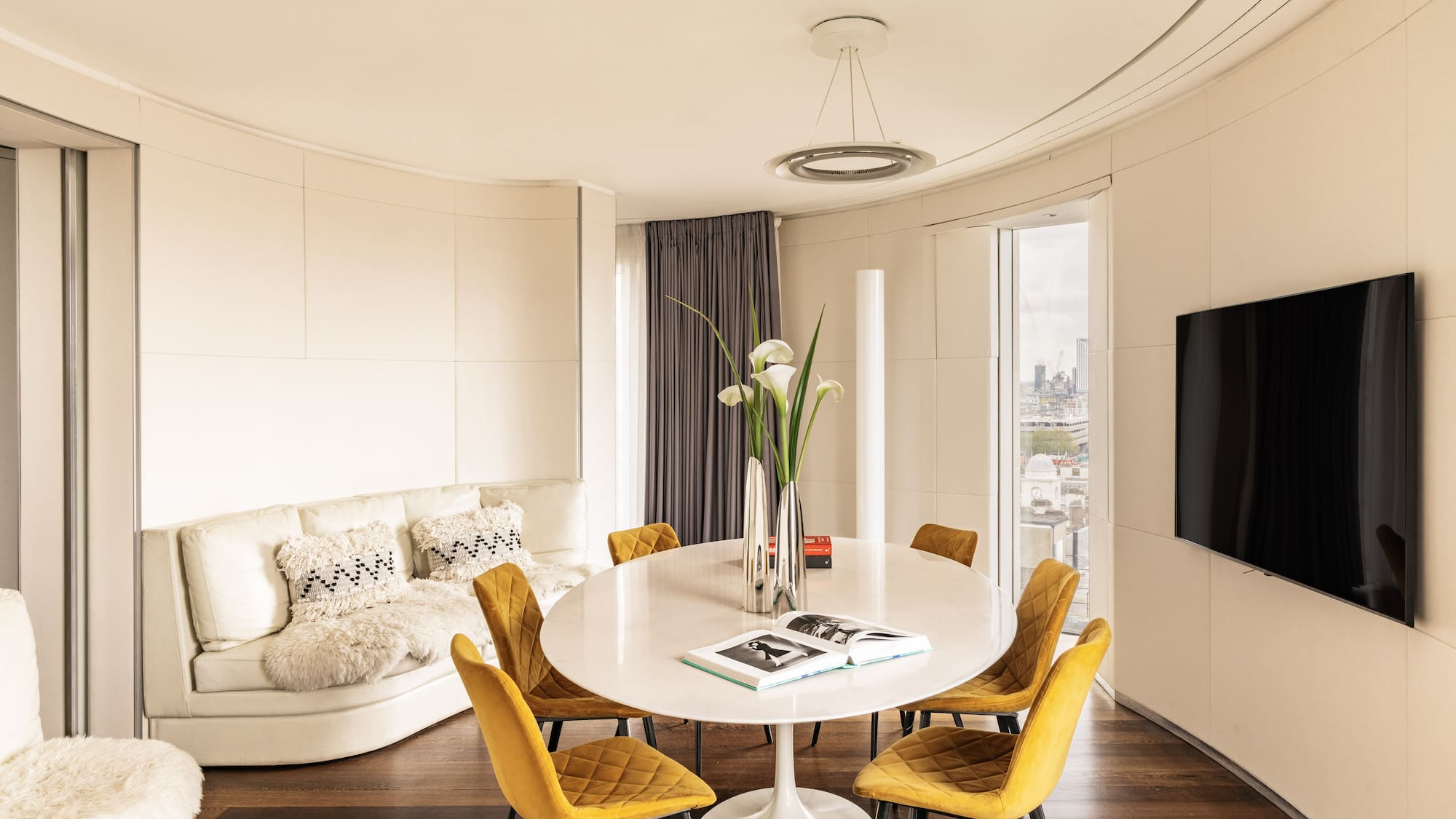 Modern living/dining room with yellow chairs, white table, curved sofa, city view.