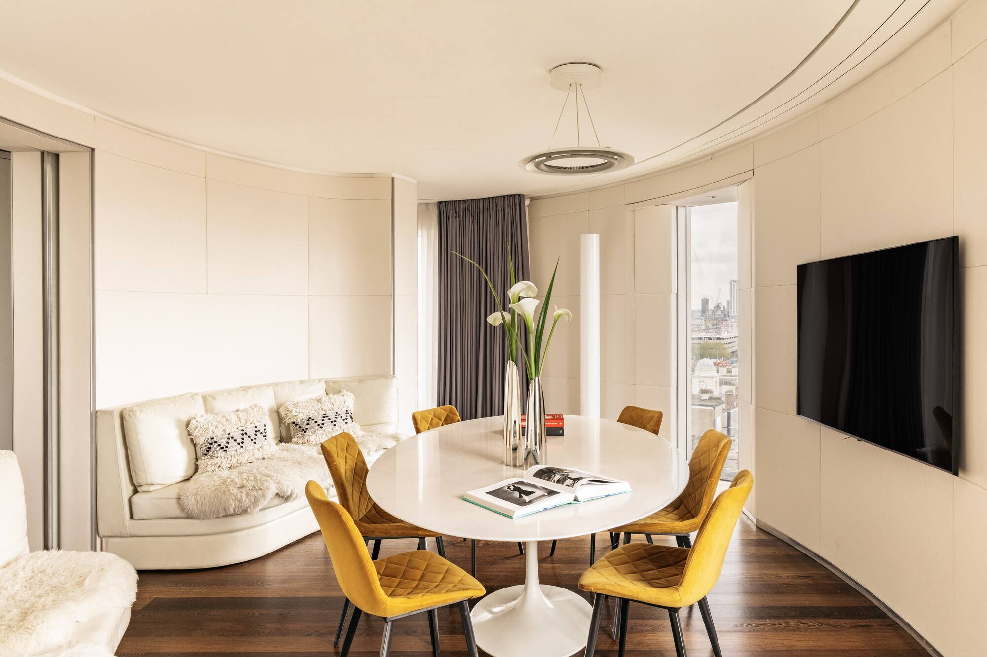 Modern living/dining room with yellow chairs, white table, curved sofa, city view.