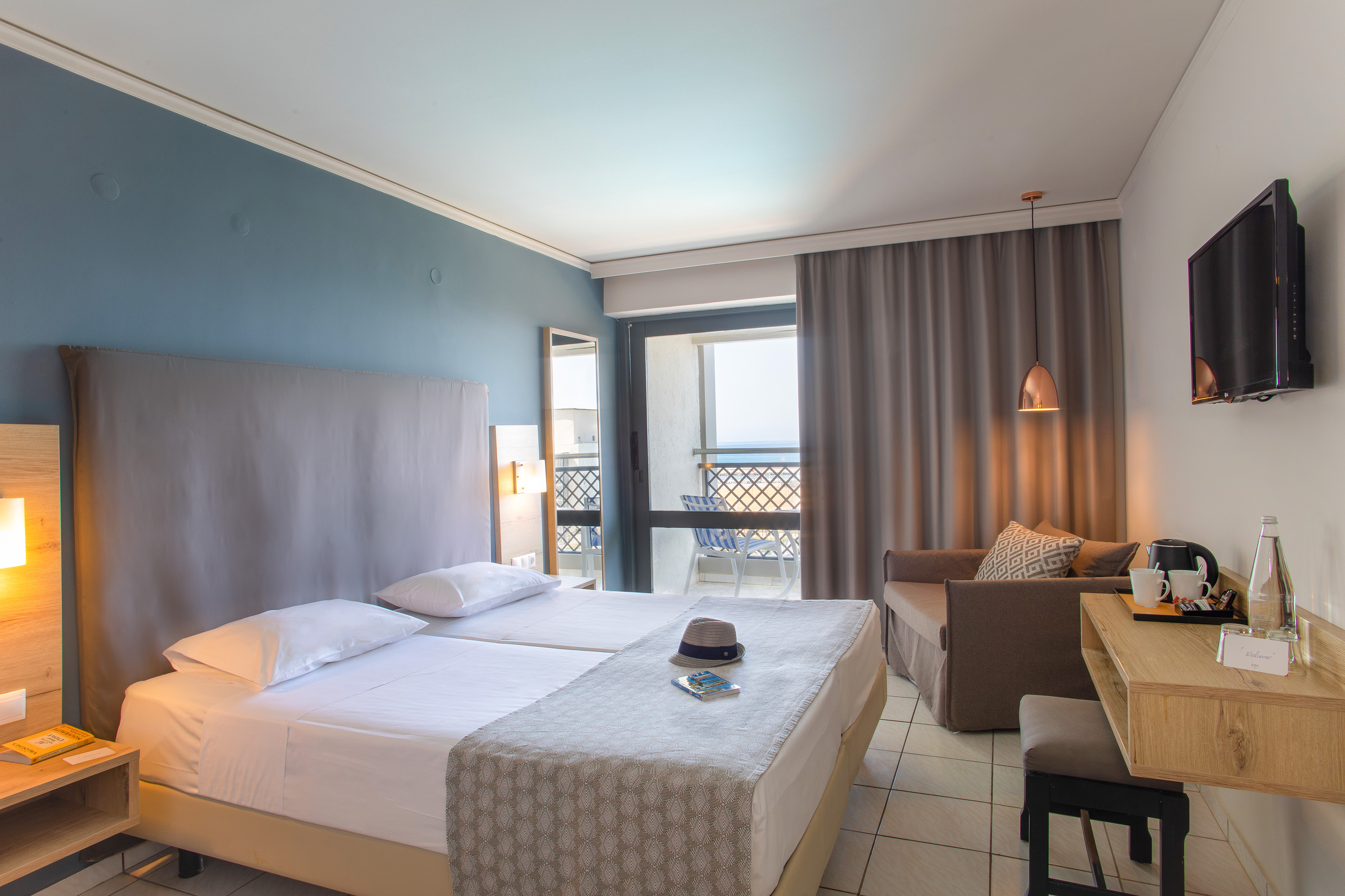 Modern hotel room with twin beds, blue wall, sofa, balcony, and sea view.