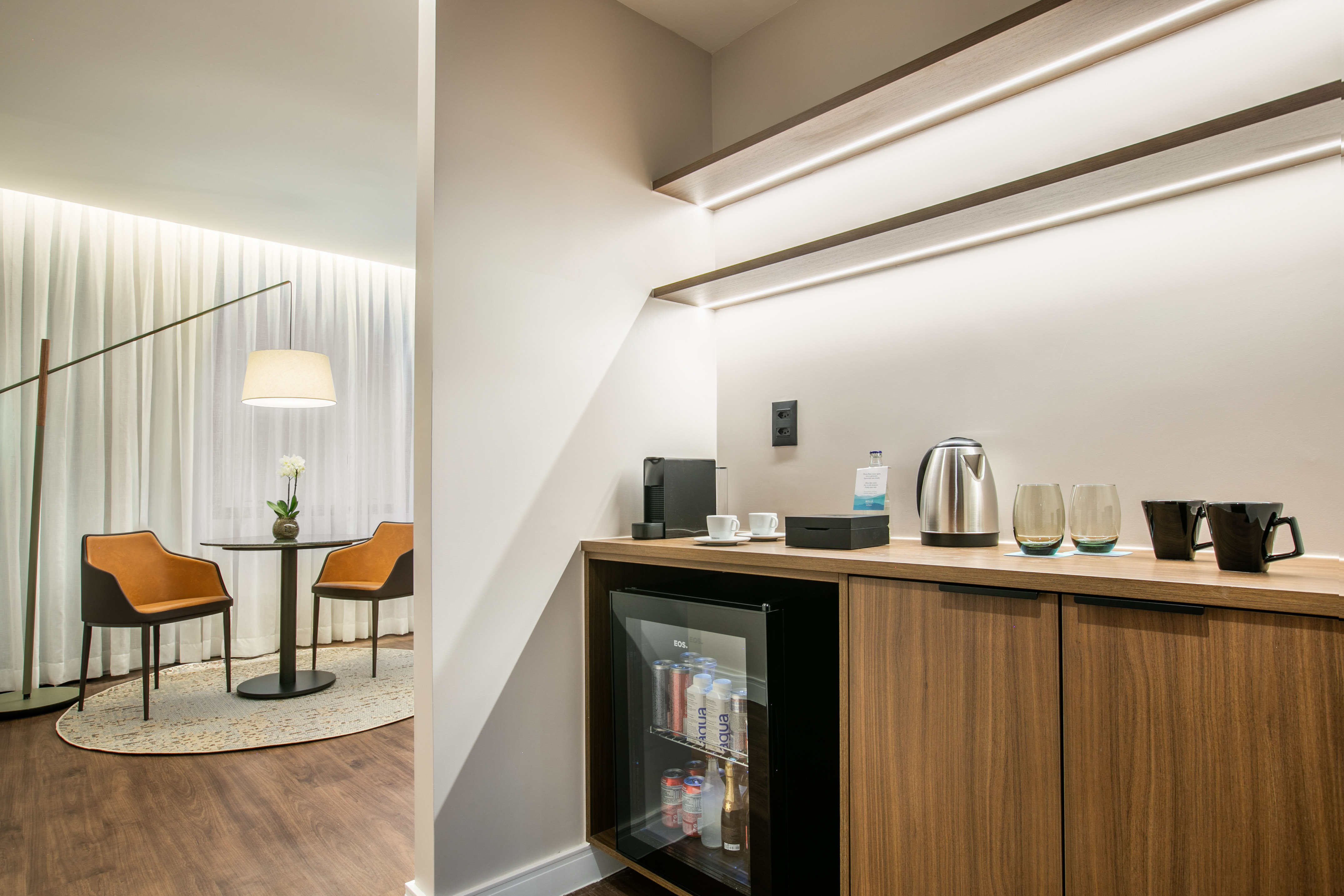 Modern hotel room with minibar, coffee machine, kettle, and cozy seating area.