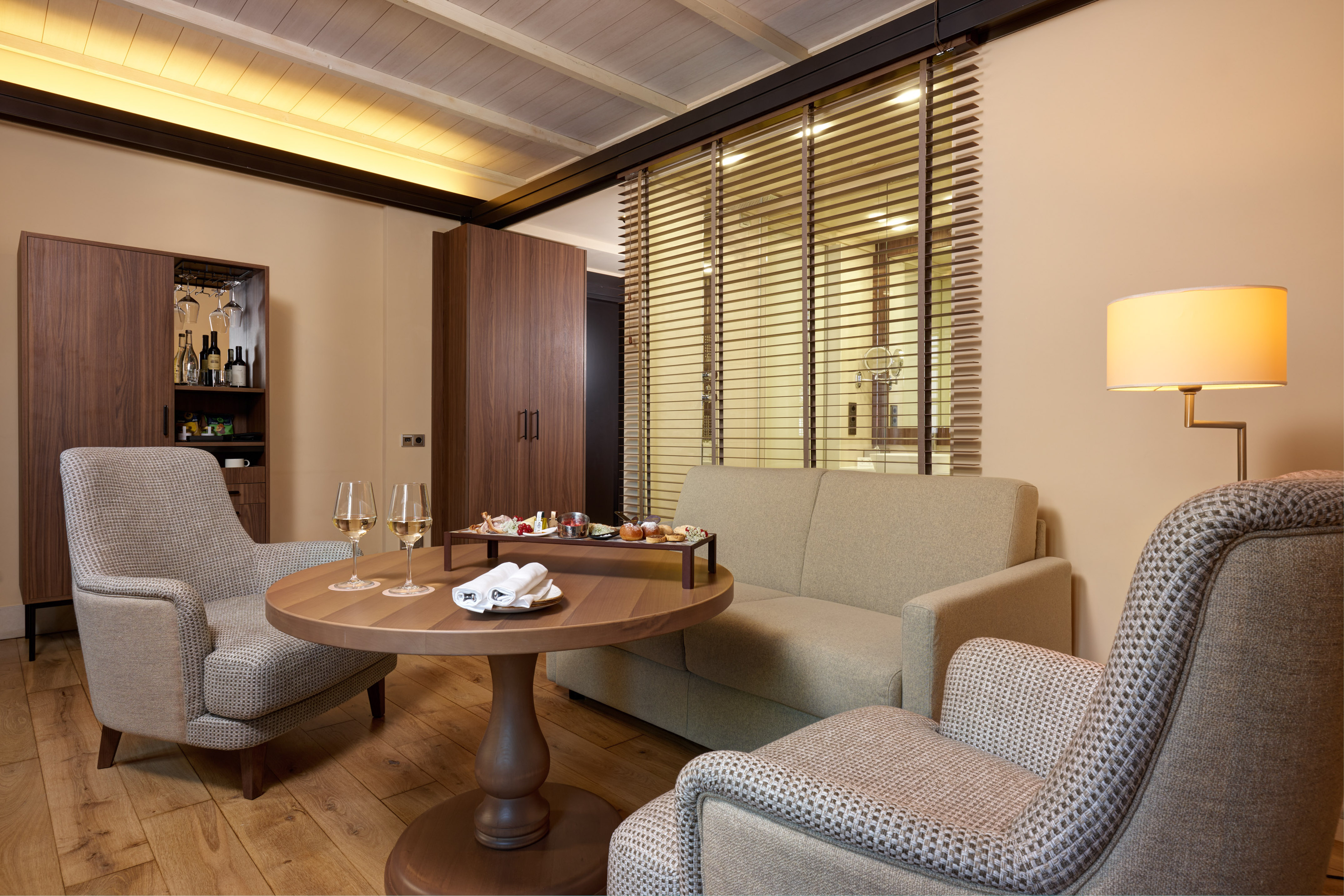 Hotel suite lounge with armchairs, sofa, wooden table, wine, snacks, and minibar.