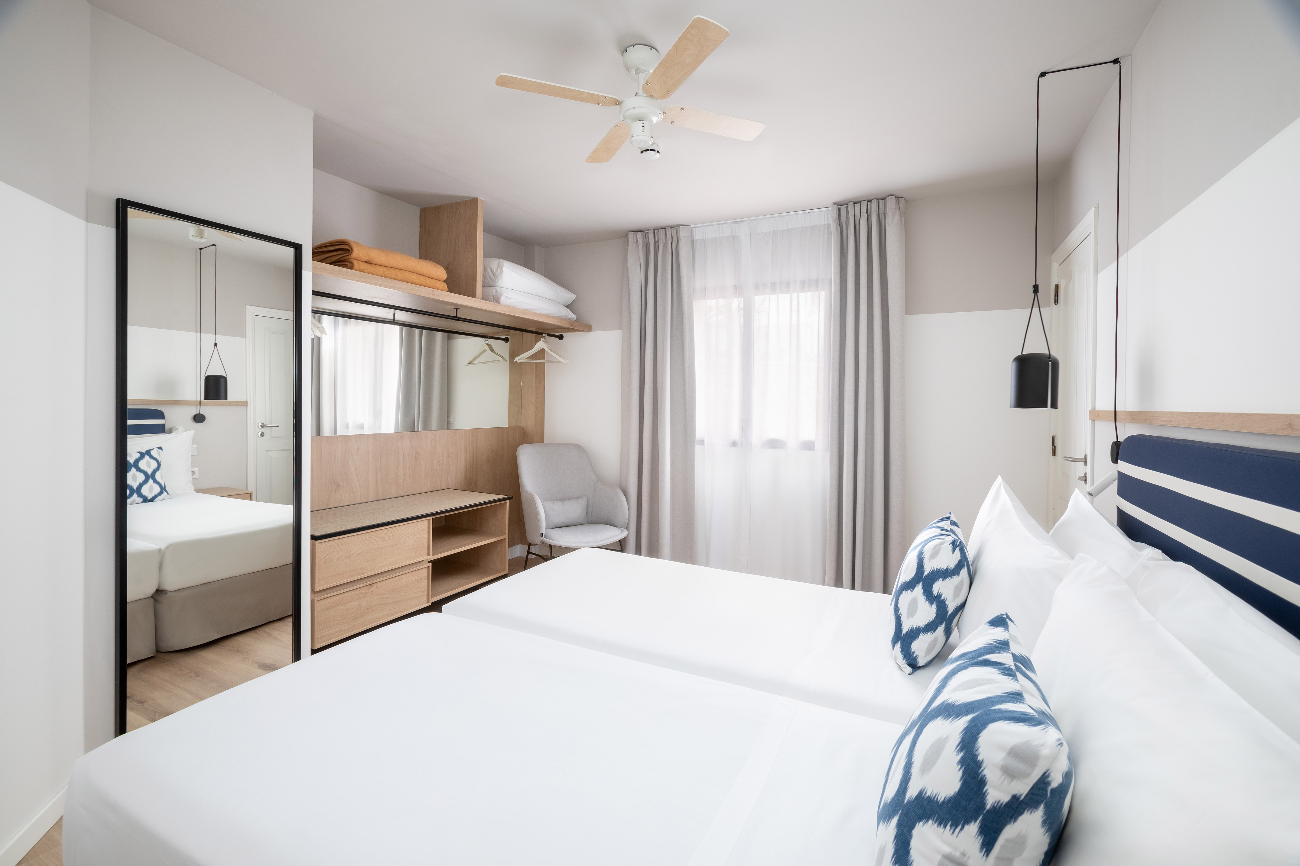 Twin hotel bedroom with white beds, blue pillows, wooden cabinet, and ceiling fan.