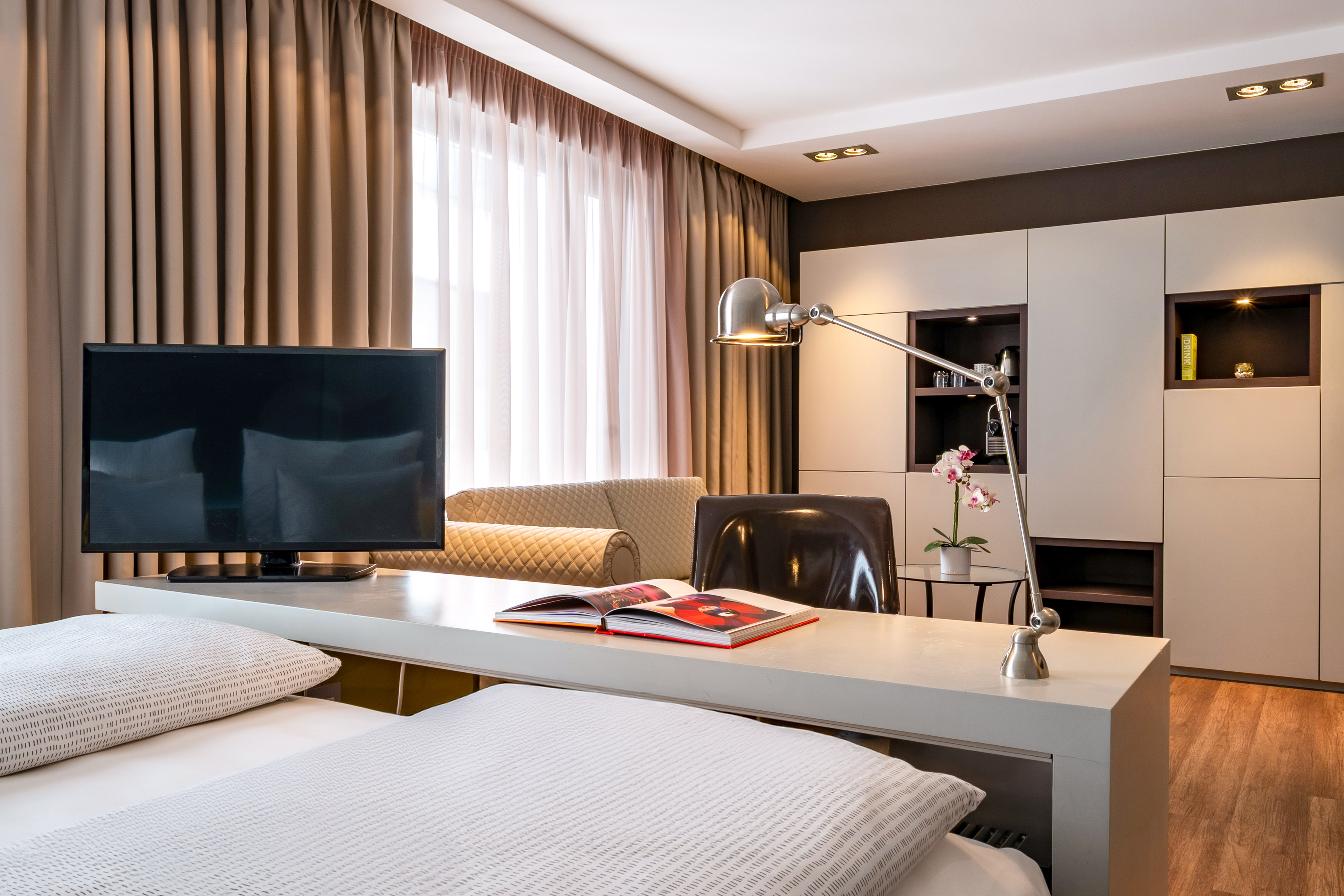 Modern hotel room with TV, desk, bed, lounge sofa, and cabinet storage.