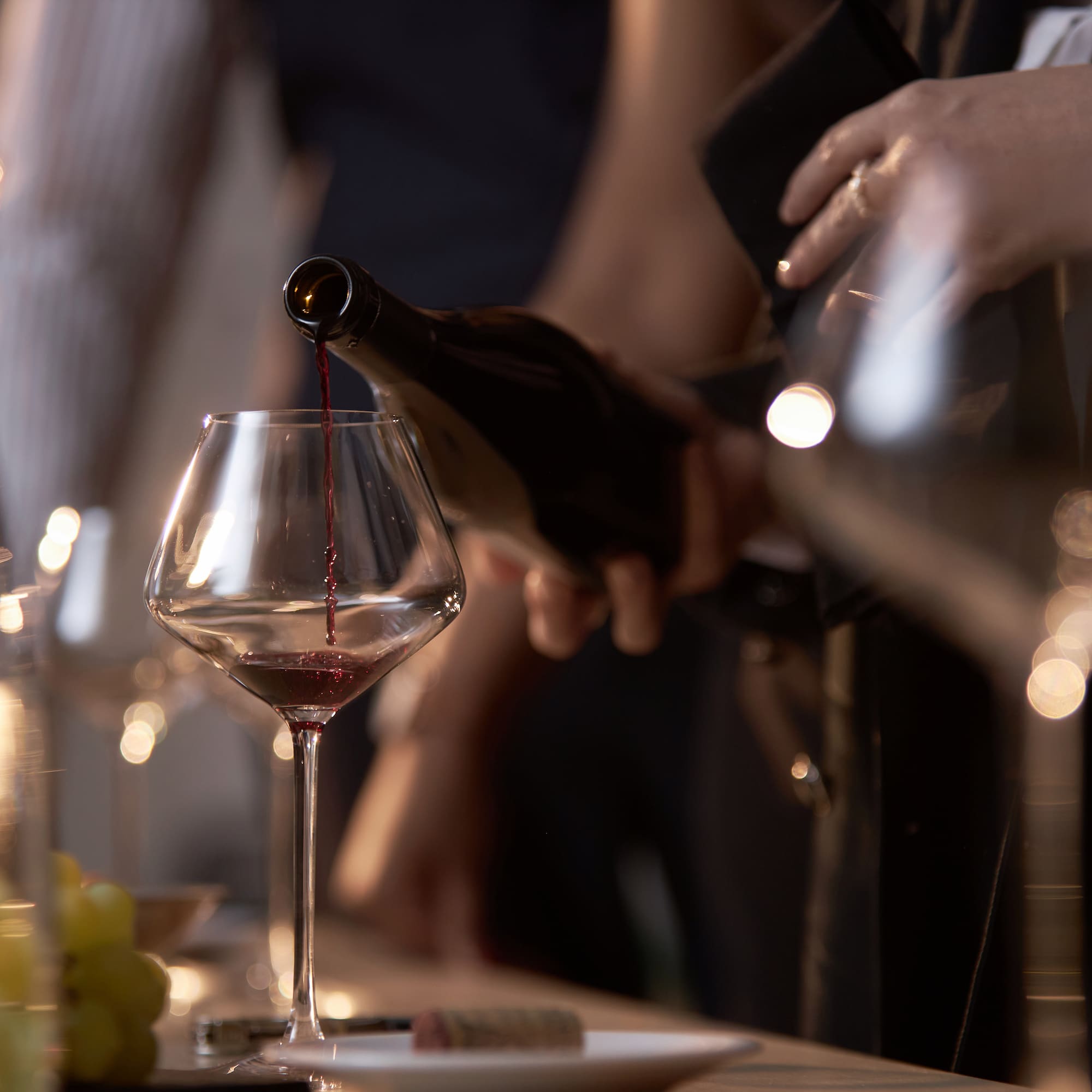 Hand pouring red wine from a bottle into a wine glass.